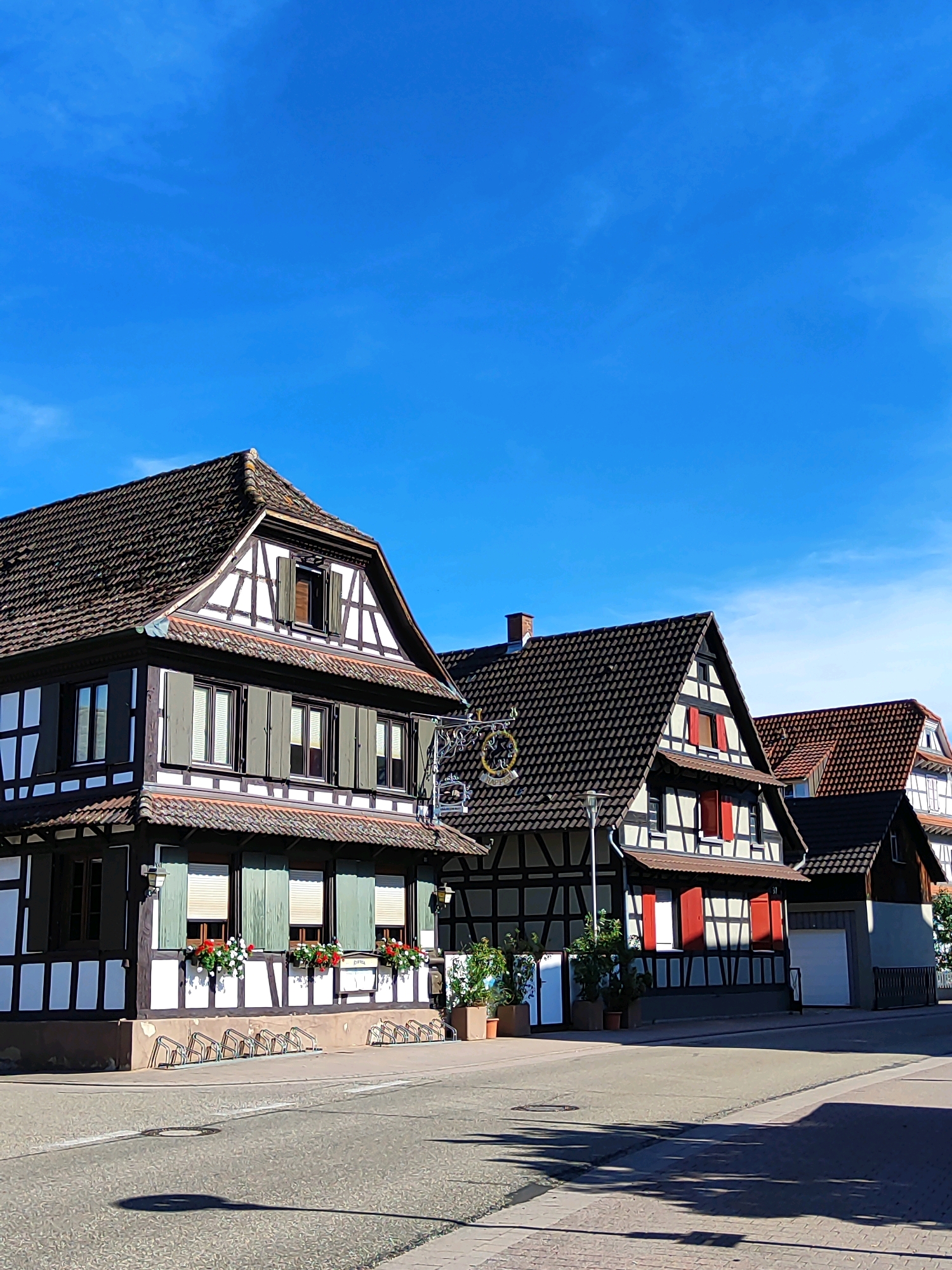 The image shows a street view in Willstätt, Germany. In the foreground, a paved road with a white lane marking runs horizontally. A circular metal manhole cover is visible on the road, casting a shadow. A shadow of a tree or pole is cast across the road and sidewalk. A row of metal bicycle racks is present on the right side of the road, in front of a building.
The buildings in the background are traditional half-timbered houses with dark tiled roofs and white plaster walls with dark wooden beams. The house on the left has dark green shutters and window boxes with red flowers. A sign with a circular emblem hangs from the building. The middle house also features half-timbering and red shutters. The house on the far right appears to be a garage with a white door. The sky is a clear, vibrant blue with a few wispy clouds. The overall scene suggests a sunny day in a German town.