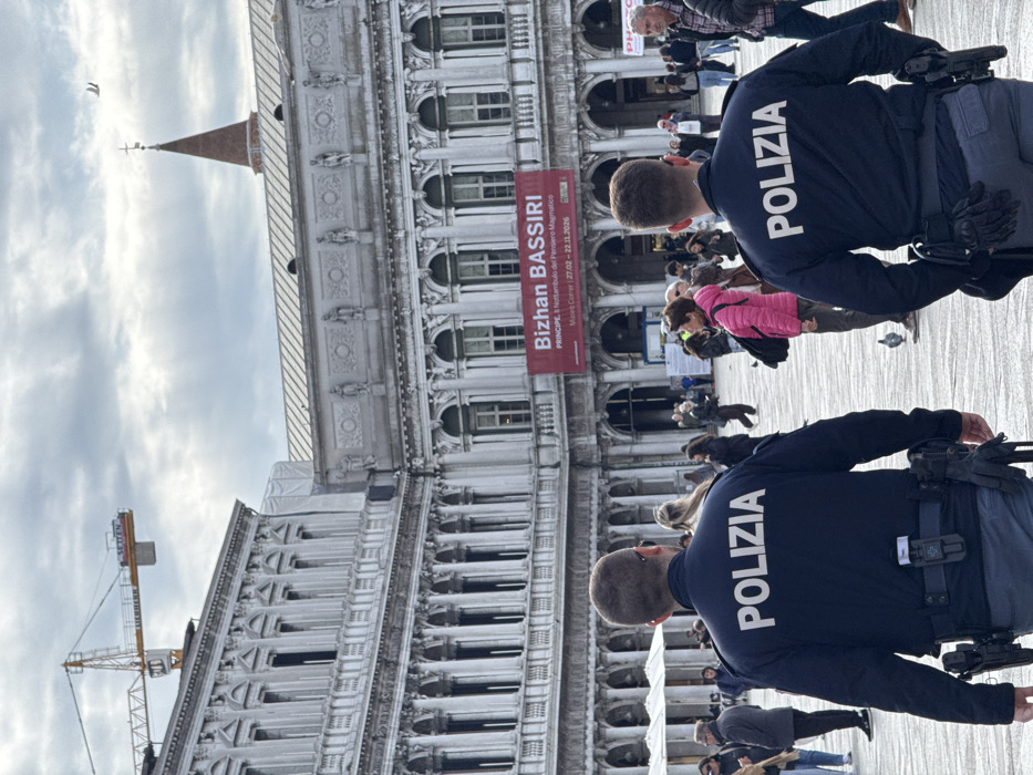 The image depicts two male Italian police officers, seen from behind, patrolling St. Mark's Square (Piazza San Marco) in Venice, Italy, under an overcast sky. Both officers wear dark blue long-sleeved shirts with "POLIZIA" emblazoned in white capital letters across the back, grey trousers, and utility belts. Each belt is equipped with a holstered firearm on the right hip and a body camera mounted on the front. The officer on the right also has black gloves tucked into his belt.
The square is populated by numerous pedestrians of diverse attire, with several pigeons visible on the paved ground. In the background, grand, ornate historical buildings, characteristic of the Procuratie, line the square. These structures feature multiple levels of arched windows, columns, and intricate stone carvings. A large red banner advertising the "Bizhan BASSIRI" exhibition at the "Museo Correr," running from "27.02 - 22.11.2026," is prominently displayed on one of the buildings. A yellow tower crane is visible above the buildings on the left, indicating nearby construction or maintenance. A reddish-brown spire topped with a cross, likely a part of St. Mark's Campanile, is visible in the upper right against the cloudy sky.