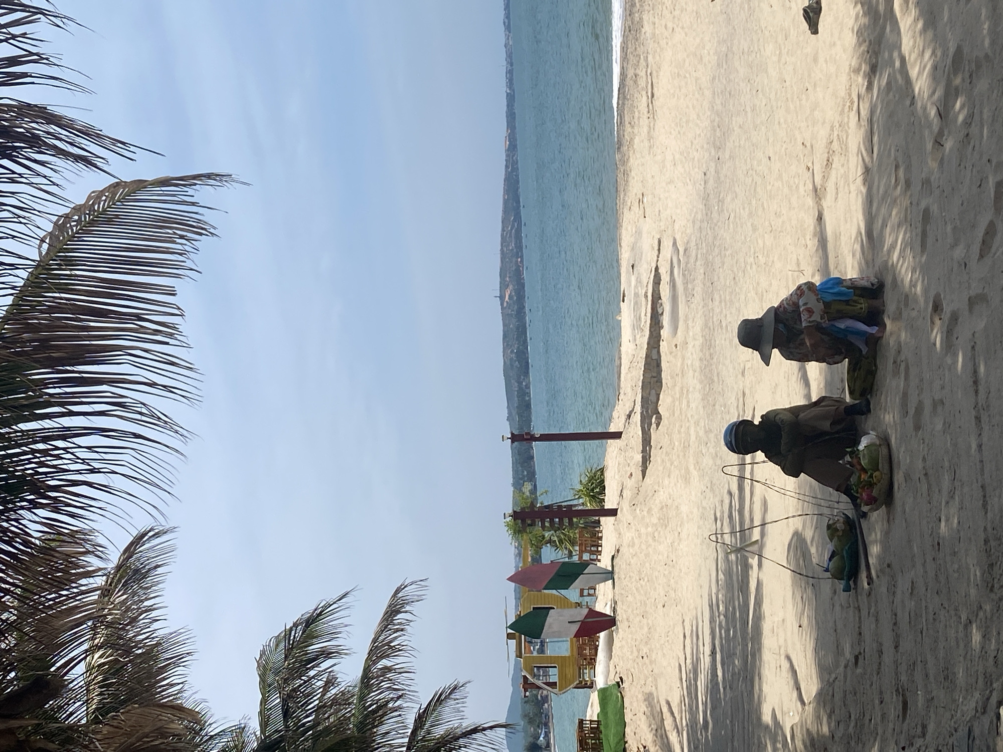 The image depicts a beach scene in Phu Thuy Ward, Vietnam, under a bright, clear sky. In the foreground, two individuals are seated on the sand, partially shaded by palm trees. The person on the left is wearing a dark top, brown pants, and a blue helmet. They are positioned next to a woven basket containing several green coconuts. To their right, a yoke-like structure with two bags hangs, also appearing to hold coconuts. The person on the right is seated and wearing a patterned long-sleeved shirt and a wide-brimmed hat, with blue and white fabric draped over their lap. A woven basket is in front of them, holding a colorful assortment of fruits.
In the mid-ground, the sandy beach extends towards the ocean. A yellow structure resembling a small helicopter is visible, with two decorative surfboards standing in front of it. One surfboard has red, white, and green stripes, while the other has pink and green stripes. Beyond these objects, wooden tables and chairs are set up on the sand. Two wooden poles with lights on top are also present on the beach.
The background features a calm, blue ocean stretching towards a distant coastline with hills and scattered buildings. A lighthouse is visible on the horizon. The water is clear, with small waves lapping at the shore. Several small boats are dotted on the sea. The overall impression is a tranquil beach environment, with local vendors present.