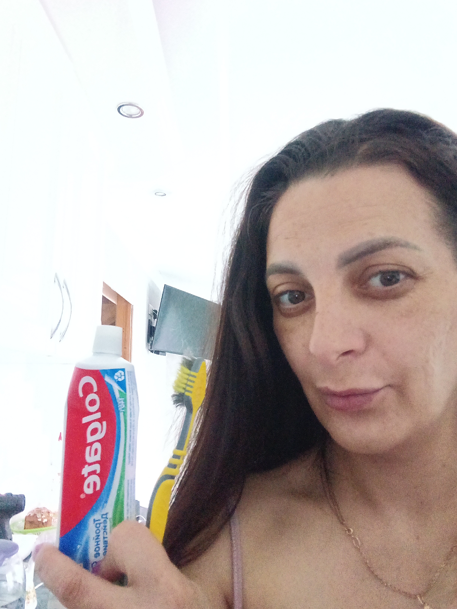 This is a selfie-style photograph of a woman indoors, possibly in a bathroom or kitchen, given the bright white walls and ceiling, and the presence of dental hygiene products. The woman, with long dark hair, looks directly into the camera with a neutral to slightly contemplative expression. She holds a tube of Colgate toothpaste in her right hand, positioned prominently in the lower-left foreground. The toothpaste tube is primarily red, white, and blue.
Visible text on the toothpaste tube appears reversed: "Colgate" is clearly legible in red. Below it, reversed Cyrillic text that appears to be "БЛЕНДЕНД" (Blended) can be discerned, along with the reversed alphanumeric code "1926A" on a green stripe. A yellow and black toothbrush is partially visible behind the toothpaste tube.
In the brightly lit background, a white ceiling features two recessed circular light fixtures. A rectangular object, possibly a small television or monitor, is mounted on a wall further back, and a wooden door frame is also partially visible. The woman wears a light-colored strap, likely from a top, and a delicate gold chain necklace around her neck. The setting is a domestic interior, consistent with the location context of Andreyevo, Russia. The bright lighting suggests it is daytime.
