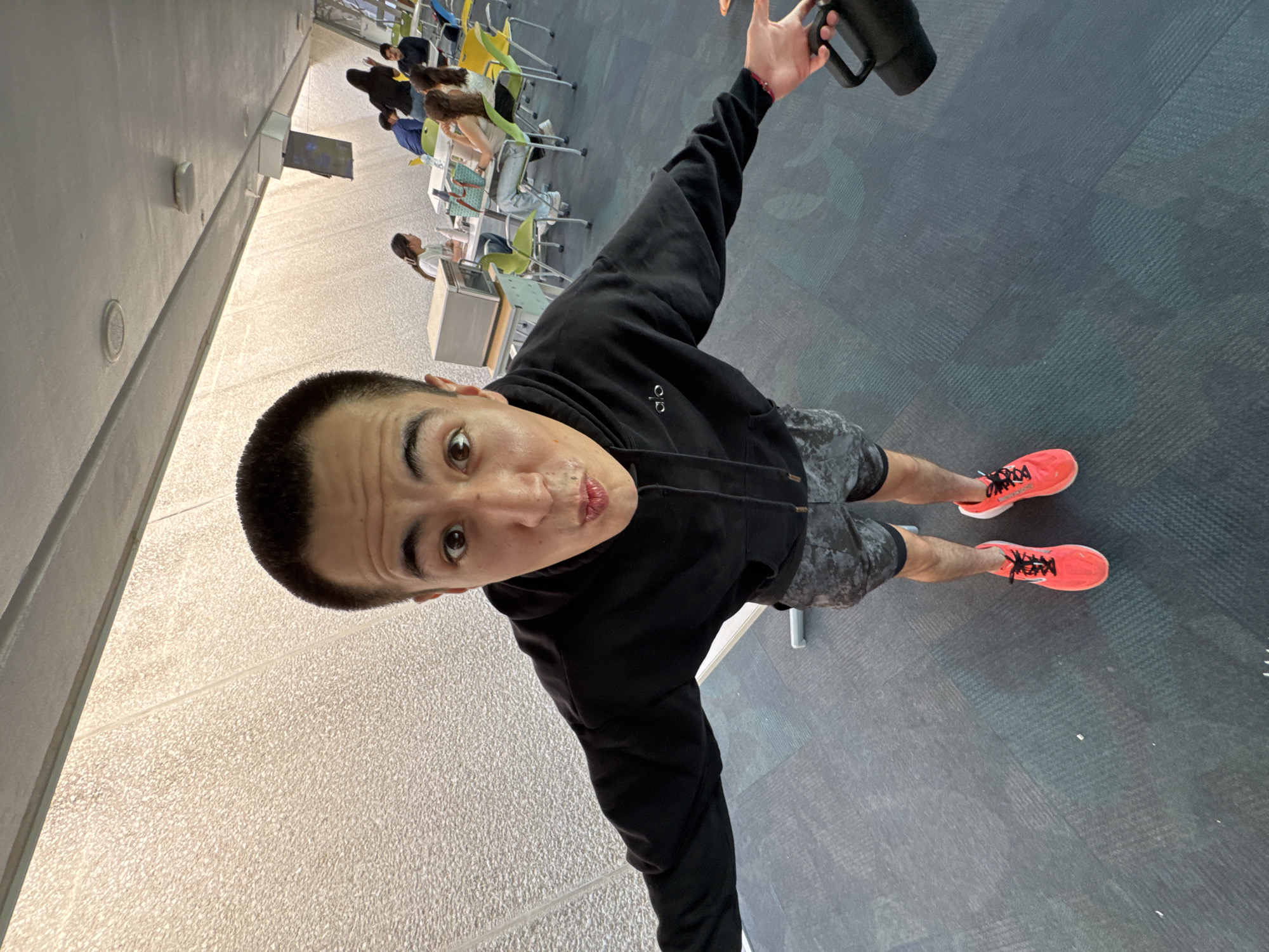 The image presents a high-angle, wide-lens shot of a young man taken indoors, likely in a public space in Puebla, Mexico. The main subject, positioned prominently, looks up towards the camera with a playful, slightly puckered lip expression. He has short, dark hair and is dressed in a black "alo" brand hoodie, camouflage-patterned shorts, and vibrant orange running shoes with black laces. He extends his right arm upwards, holding a black mug or insulated bottle.
The setting appears to be a cafeteria, food court, or communal study area. The floor beneath the man is covered with a dark gray patterned carpet. To his left, a light-colored wall with visible architectural details like recessed lights and vents rises. In the background, several people are seated at various tables with colorful chairs (yellow, green, blue). These individuals are engaged in typical activities such as eating, socializing, or using electronic devices. A white counter or serving station and a wall-mounted screen are also visible further back.
The overall scene suggests a casual daytime moment, with ample ambient lighting. The man's expressive pose indicates a selfie or a candid, deliberately composed photograph.