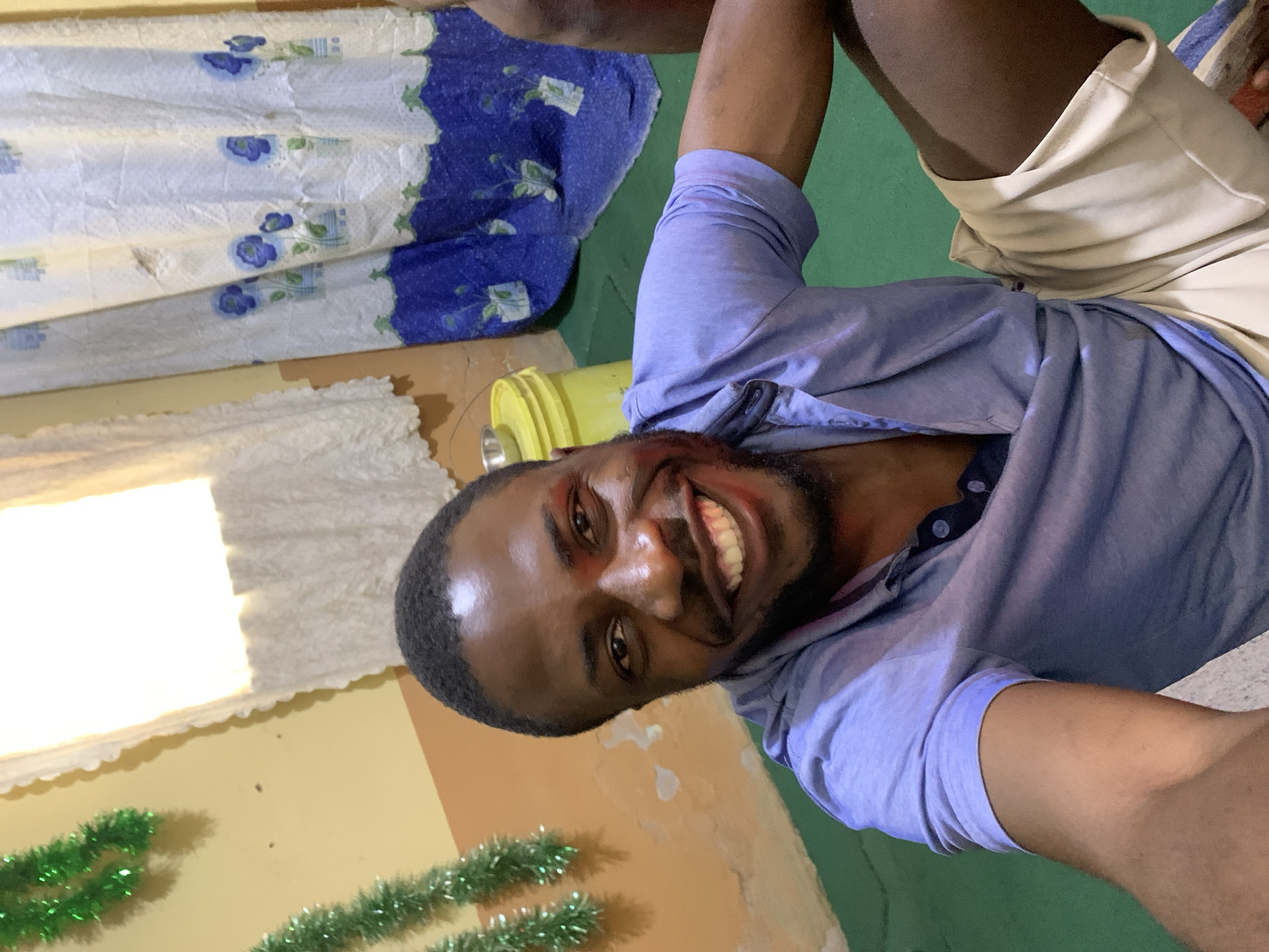 A man with dark skin and short black hair is pictured from the chest up, smiling directly into the camera. He is wearing a light blue short-sleeved polo shirt with two dark blue buttons at the collar and light beige shorts with a blue stripe down the side. His left arm is bent, resting on his lap. Behind him, the background consists of a pale yellow wall with some textured sections, a green floor, and to the right, a blue and white curtain with a floral pattern. Green garland is visible on the left side of the image, suggesting a festive decoration. A yellow bucket with a metal bowl on top is also visible to the left of the curtain. The lighting suggests an indoor setting, possibly a home, with natural light coming from a window behind the white lace curtains. The overall scene appears to be informal. The man's attire and the background suggest an everyday setting. The city of Rafin Par, Nigeria, is mentioned as the location, but no specific landmarks or identifying features of the city are visible in the image.