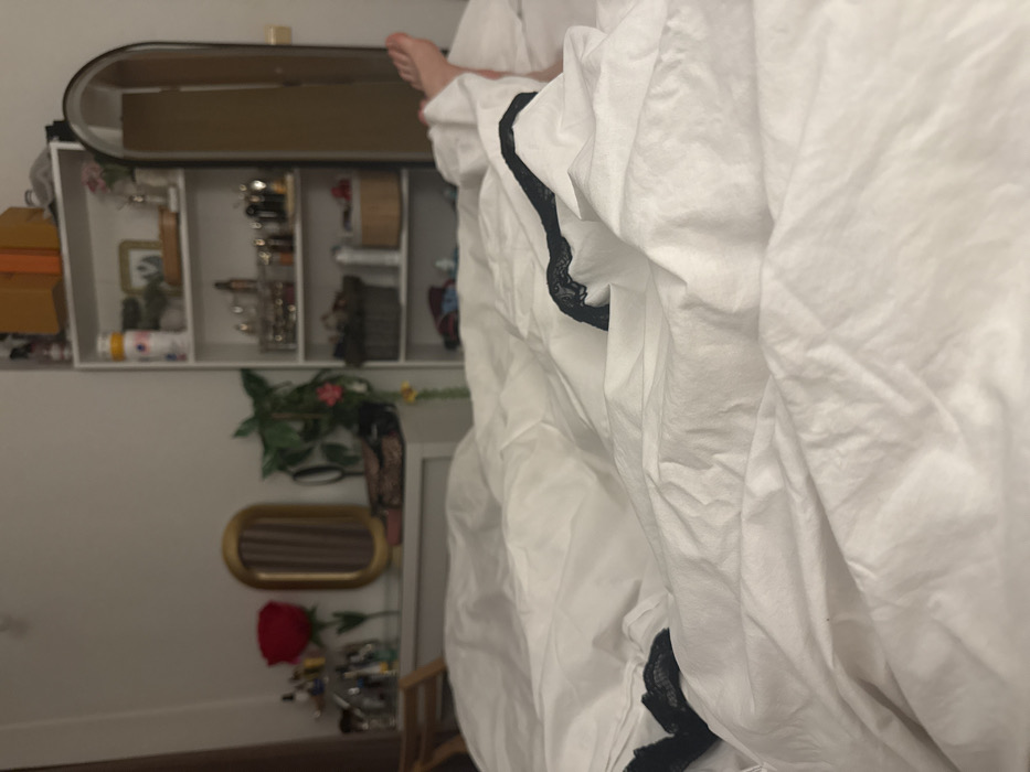 The image depicts a residential bedroom interior in Sofia, Bulgaria, viewed from a low perspective at the foot of a bed. The foreground is dominated by crumpled white bedding, within which a person's bare feet are partially visible towards the right side. A black, lacy garment, possibly lingerie, is partially uncovered by the white sheets on the left and right sides of the bed, suggesting it is on or near the occupant.
The background, partially out of focus, reveals a room furnished with personal items. To the left, a light-colored dresser or vanity surface displays a collection of cosmetic bottles and containers, a single red rose in a vase, an oval gold-framed mirror, a small green potted plant, and a patterned bag. To the right, a tall, white open shelving unit contains multiple tiers of objects, including an orange box, various bottles, a small framed picture, an acrylic organizer filled with cosmetic products, a dark wicker basket, and folded garments on the bottom shelf. Adjacent to the shelving unit stands a tall, arched, full-length mirror reflecting a light-colored door. The overall lighting is dim, indicating an indoor setting, possibly evening or low ambient light conditions.