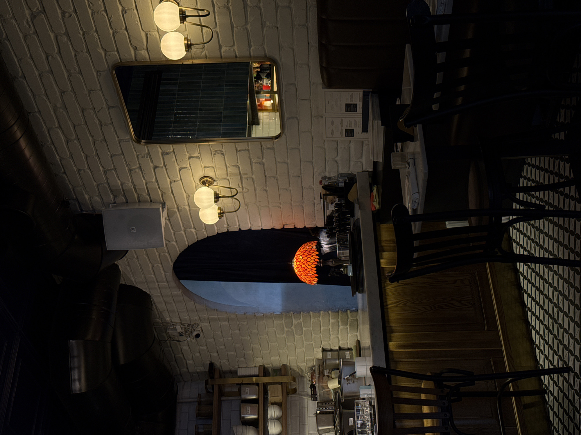 The image shows the interior of a dimly lit establishment, possibly a cafe or restaurant, in Yerevan, Armenia. The walls are covered in white brickwork, and the ceiling features exposed dark metal pipes and an arched opening. A rectangular mirror with a gold frame hangs on the brick wall, reflecting a dimly lit area with shelves and bottles.
Several light fixtures are visible: two globe-shaped lights mounted on a gold fixture above the mirror, and another similar fixture with two globe lights below it. An ornate, orange, stained-glass pendant light hangs in the arched opening.
The scene includes several dark wooden chairs with slatted backs, some of which are pulled up to wooden tables. In the background, shelves are stocked with various items, including what appear to be containers and possibly beverages. A white speaker is mounted on the wall. The overall ambiance is moody and intimate, suggesting it is evening. There is no visible text in the image.