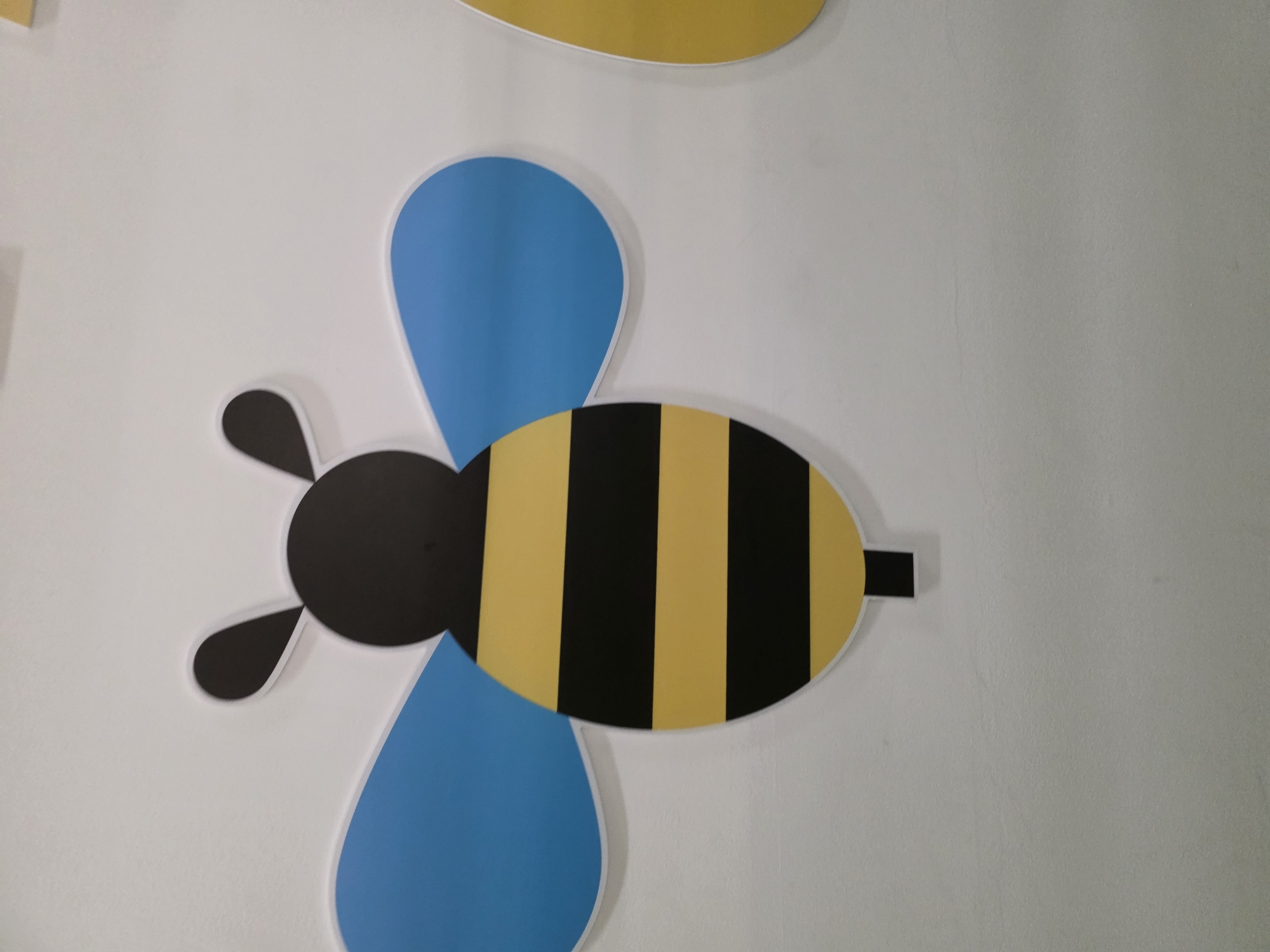 A stylized, cartoon bee decoration is affixed to a plain white wall. The bee has a black head with two antennae. Its body is oval-shaped and features horizontal stripes of yellow and black. Two large, blue, teardrop-shaped wings extend from the sides of its body. A small, black rectangle extends downwards from the bottom of the bee's body, suggesting legs or a stinger. The lighting is even, and the wall appears to be indoors, with no visible windows or doors.
