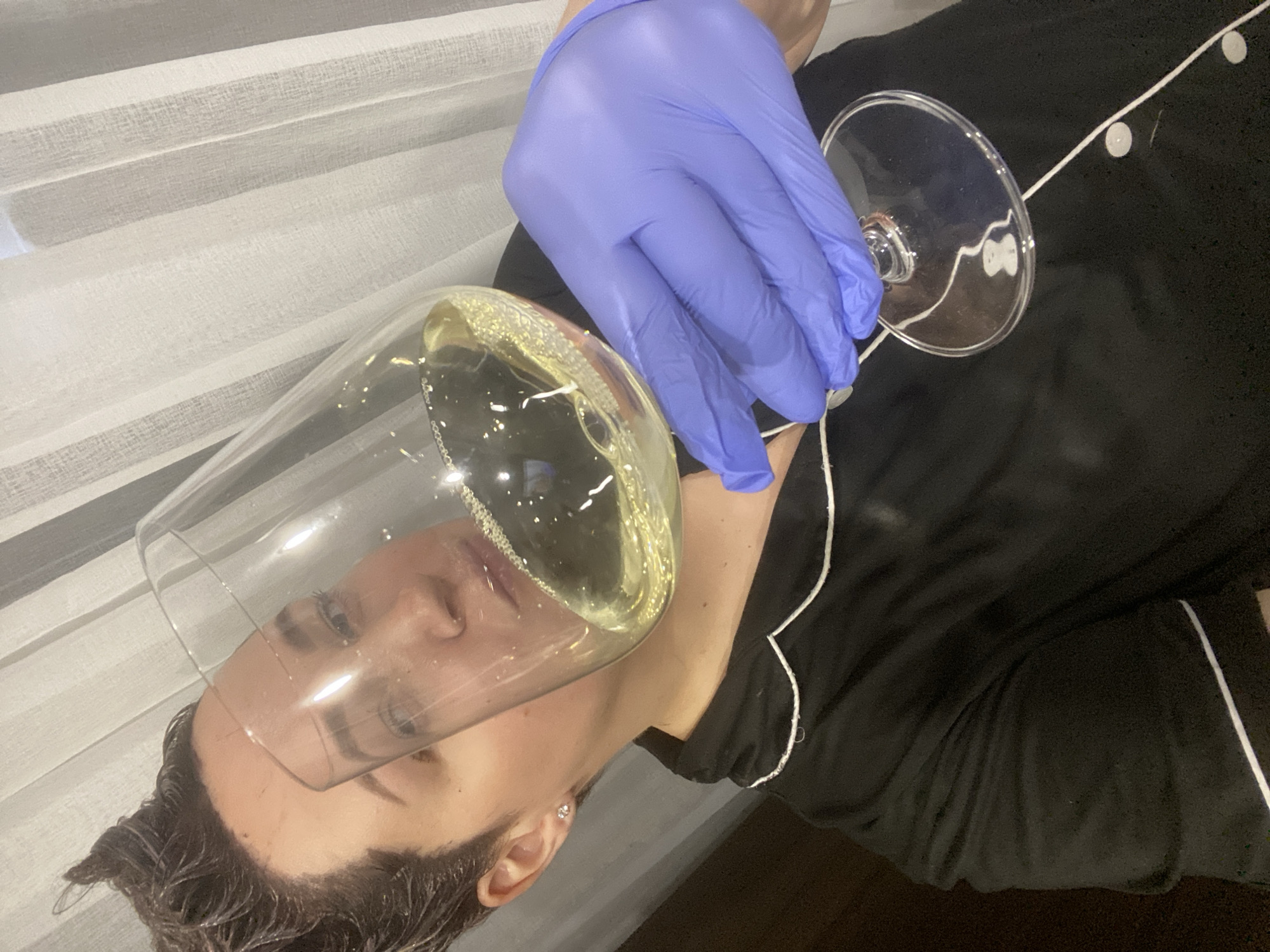 The image captures a close-up, rotated view of a person lying down indoors, engaged in an unusual interaction with a stemmed glass.
**Subjects:** The primary subject is a person, likely a woman, visible from the chest upwards. Their dark, possibly damp hair is styled to the side, and one ear with a small stud earring is visible. A portion of their face, including their eyes, is seen through the transparent glass. A hand wearing a distinct purple latex or nitrile glove holds the stem of a large wine or champagne glass. The glass is filled with a sparkling, yellowish liquid, possibly champagne or a similar beverage, containing small bubbles and what appears to be glitter or reflective particles. The person is dressed in a black garment, resembling pajamas or loungewear, with contrasting white piping along the seams. A white string-like object, possibly decorative lights, with small white circular elements, is draped across the black fabric near the base of the glass.
**Activity or event:** The gloved hand is holding the glass with the liquid precariously positioned directly above the person's face. This setup suggests a deliberate, possibly playful or artistic act, such as preparing for a photo shoot or a theatrical moment where the liquid might be poured or spilled onto the face.
**Notable details:** The scene takes place indoors, with a backdrop of white, translucent curtains or drapes providing a soft, diffused lighting environment. The image is oriented sideways (rotated approximately 90 degrees counter-clockwise). The person's expression, viewed through the glass, appears neutral, looking upwards. The specific location of Sochi, Russia is provided, though no visual cues within the image confirm this. No visible text is present in the image.
