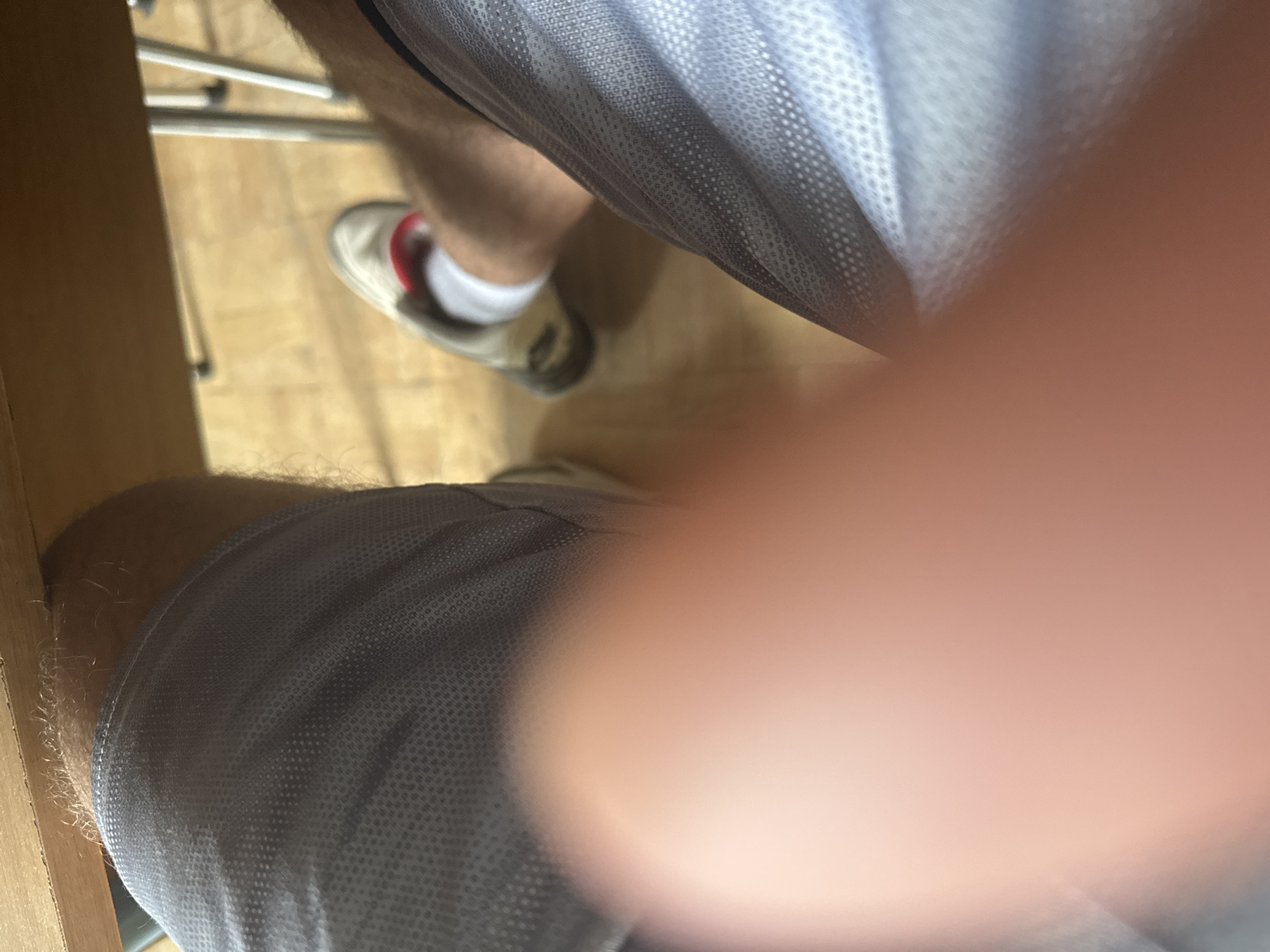 The image shows a person's legs and feet, partially obscured by a hand or finger in the foreground. The person is wearing light grey athletic shorts and white socks with a red band around the ankle. One foot is visible, wearing a light-colored sneaker with dark soles and a red accent. The person is sitting under a wooden surface, possibly a table or desk, with metal chair legs visible in the background. The floor appears to be made of light-colored wood or laminate. The scene suggests an indoor setting, possibly a home or office, in Sofia, Bulgaria.