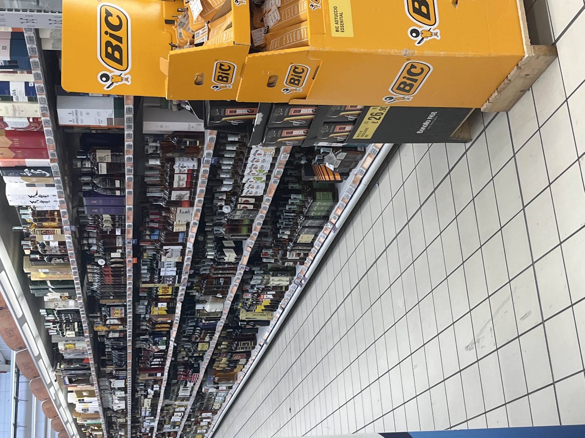 The image shows a supermarket aisle with shelves stocked with various alcoholic beverages. In the foreground, there is a bright yellow display stand with "BiC" branding, promoting what appear to be lighters or stationery products. The shelves are filled with bottles of wine, spirits, and other liquors. The flooring is tiled with a white and grey checkered pattern. In the background, other aisles of the supermarket are visible. The scene is well-lit, typical of a retail environment. The specific location in Castiglione delle Stiviere, Italy, is not evident from the image itself but is provided as context. There are no people visible in the image.