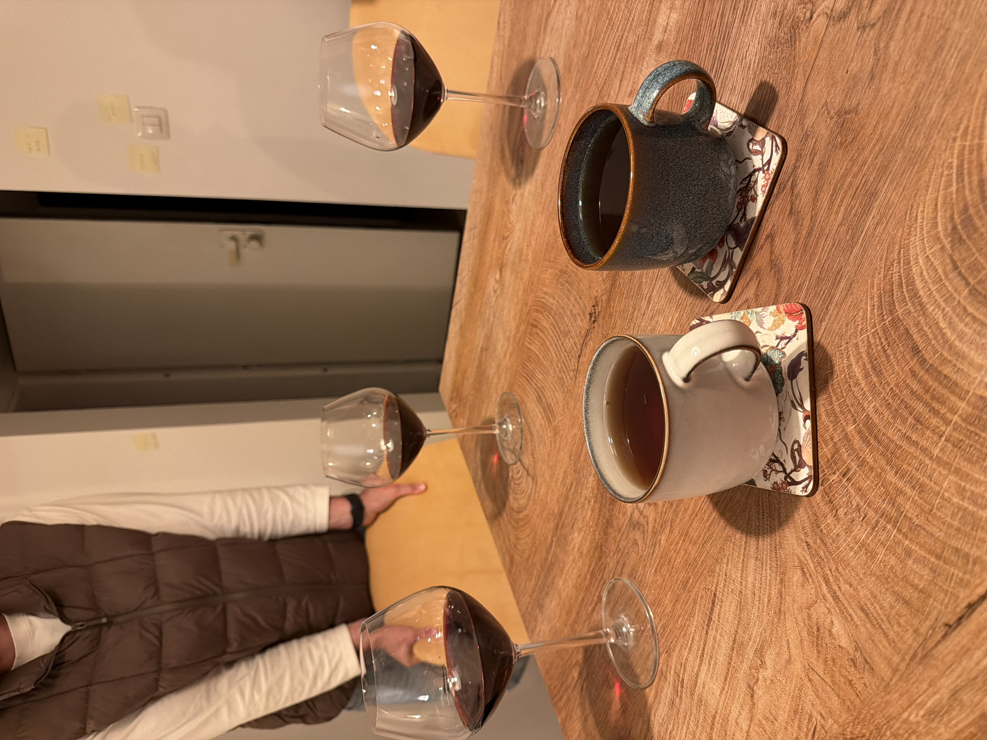 The image captures an indoor, intimate setting, likely a casual gathering around a wooden table. The central subject is a thick, rustic wooden table, featuring prominent wood grain and growth rings. On the table, there are five beverages: three wine glasses filled with red wine, and two ceramic mugs resting on decorative, floral-patterned coasters, each containing a dark, hot beverage such as coffee or tea. One mug is dark blue-grey, the other light grey-white.
A person is partially visible on the left side of the frame, wearing a white long-sleeved shirt and a brown quilted vest. Their hands are visible near a wine glass, suggesting they are participating in the gathering. The background on the left consists of a light-colored wall or cabinet. On this surface, three small, square yellow sticky notes are attached, though the text on them is illegible. Near the sticky notes, there's a small, square, clear object, possibly a light switch or hook. Below this, there's a darker horizontal line, perhaps a counter edge, and a lighter panel with a small, indistinct latch or handle.
The lighting is warm and artificial, indicating an indoor setting, likely in the evening or at night. The overall atmosphere appears relaxed and social. The location context is Rishon LeZion, Israel, though no specific visual cues within the image confirm this.