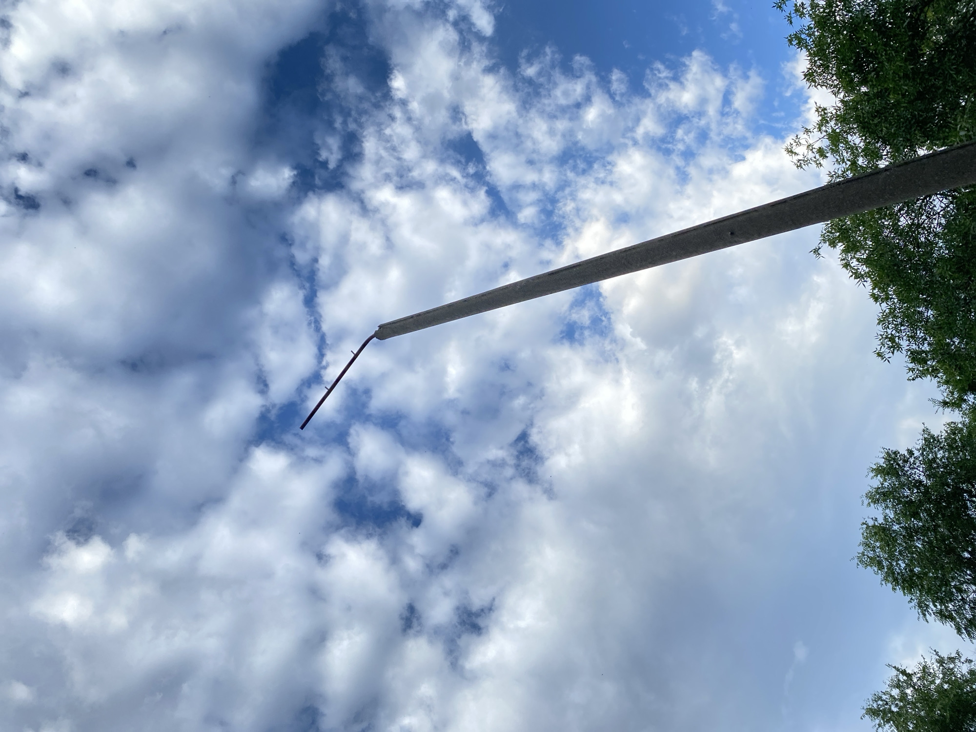 The image shows a low-angle view of a cloudy blue sky with a concrete pole extending diagonally across the frame from the left to the right. The pole appears to be a utility pole or a lamppost. A thin, dark, rod-like object is attached to the end of the pole, extending further into the sky. Trees with green foliage are visible along the right edge of the image, framing the sky and the pole. The sky is filled with a mix of white and gray clouds, indicating partly cloudy weather. The lighting suggests it is daytime, possibly late afternoon, given the slight haziness and diffused sunlight. There is no visible text in the image. The scene evokes a sense of looking upwards into the vastness of the sky, with man-made structures and nature coexisting.