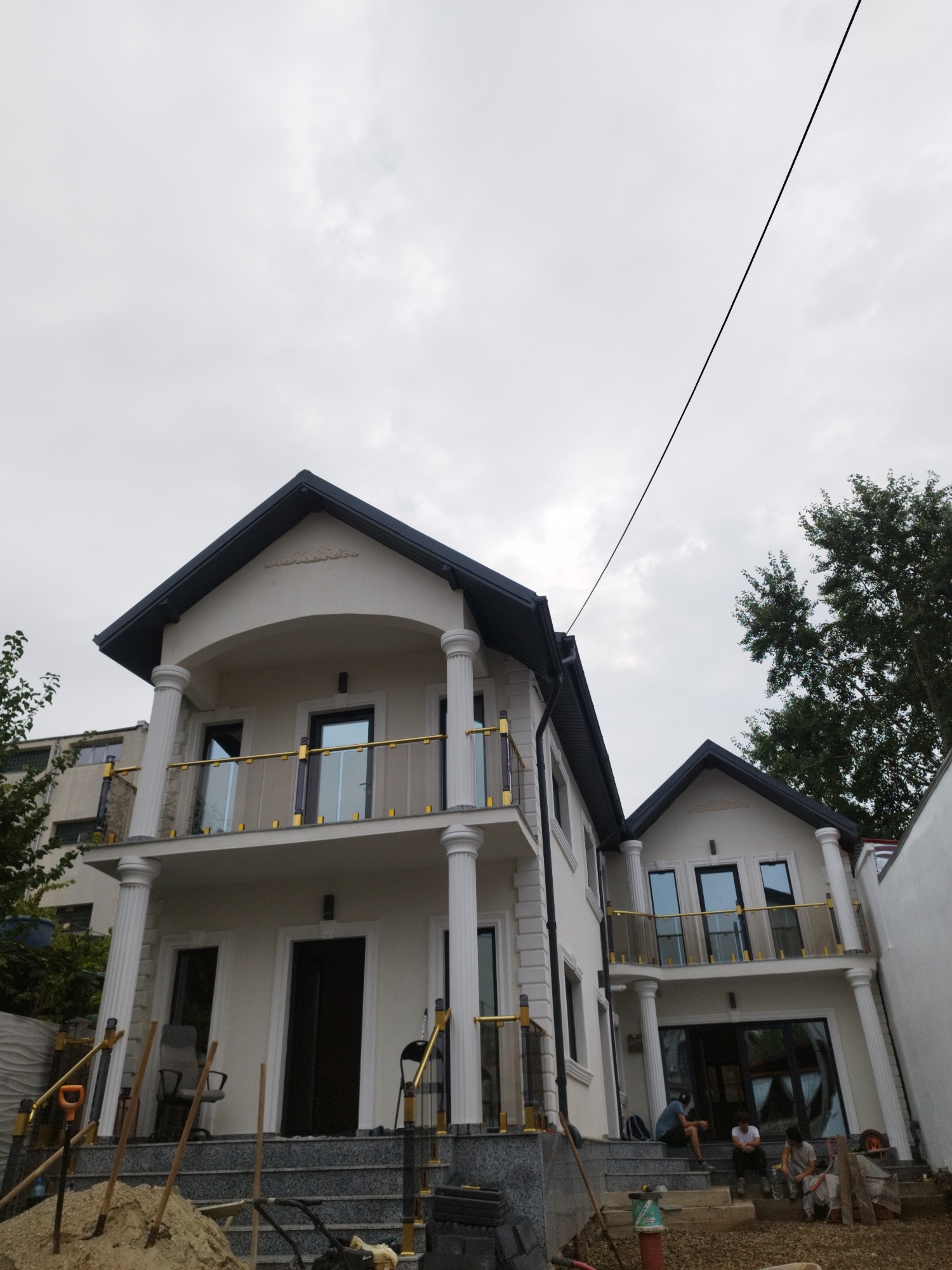 The image shows a two-story house under construction in Bucharest, Romania. The house has a modern design with a light-colored facade, dark roofing, and large windows. There are balconies on both floors with glass railings and gold accents. In front of the house, there is a set of stone stairs leading to the entrance. Construction materials, including sand, shovels, and some tools, are visible on the ground. Three individuals are seated on the stairs, seemingly taking a break. The sky is overcast, suggesting cloudy weather. The surrounding area appears to be a residential neighborhood with other buildings and trees visible in the background.