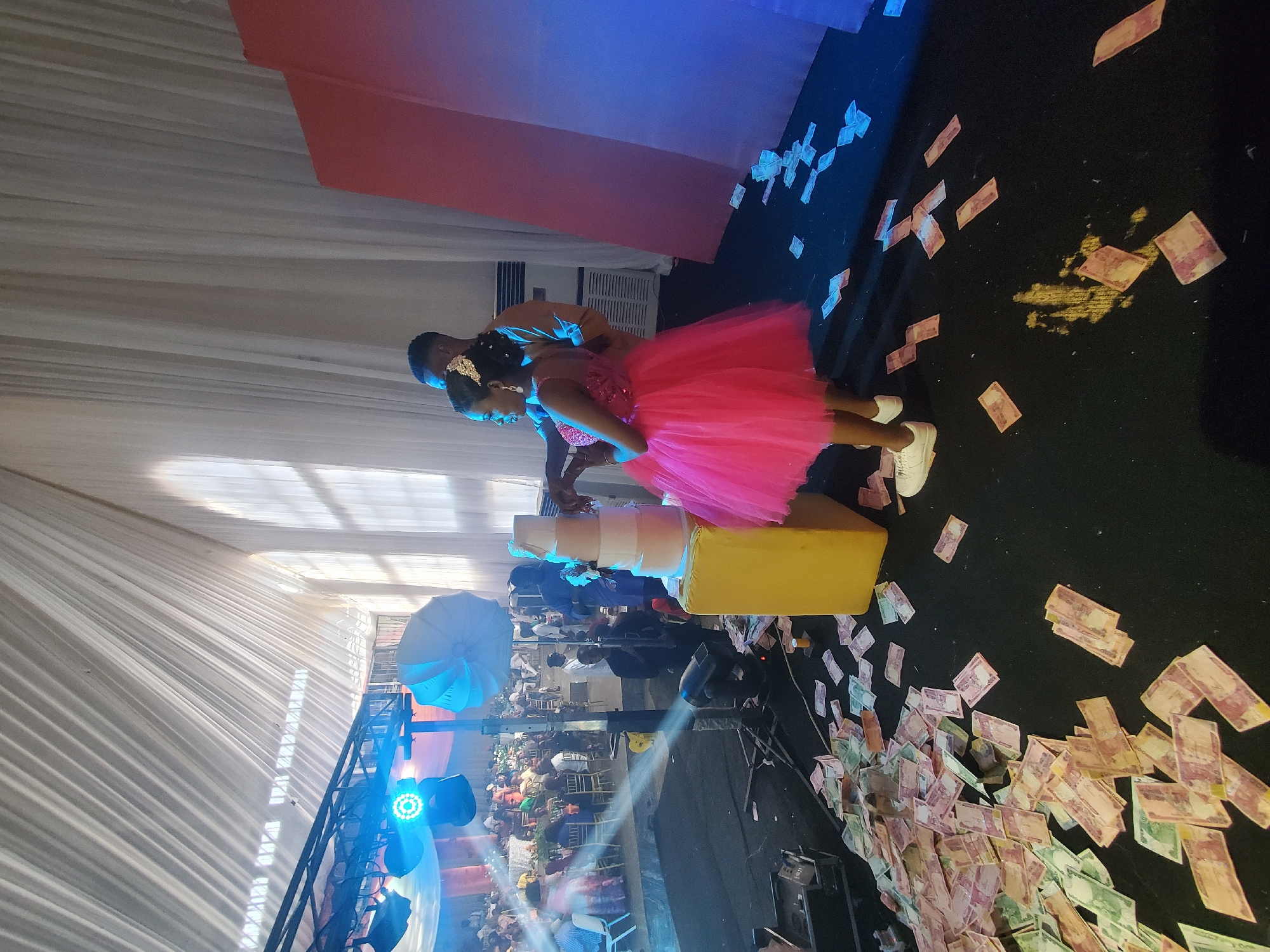 The image depicts a social gathering, possibly a wedding or celebration, in an indoor venue within the city of Unknown, Unknown. A young woman in a vibrant pink dress stands next to a multi-tiered cake, assisted by a man behind her. The floor around them is scattered with banknotes. A crowd of people is visible in the background, along with stage lighting and equipment.