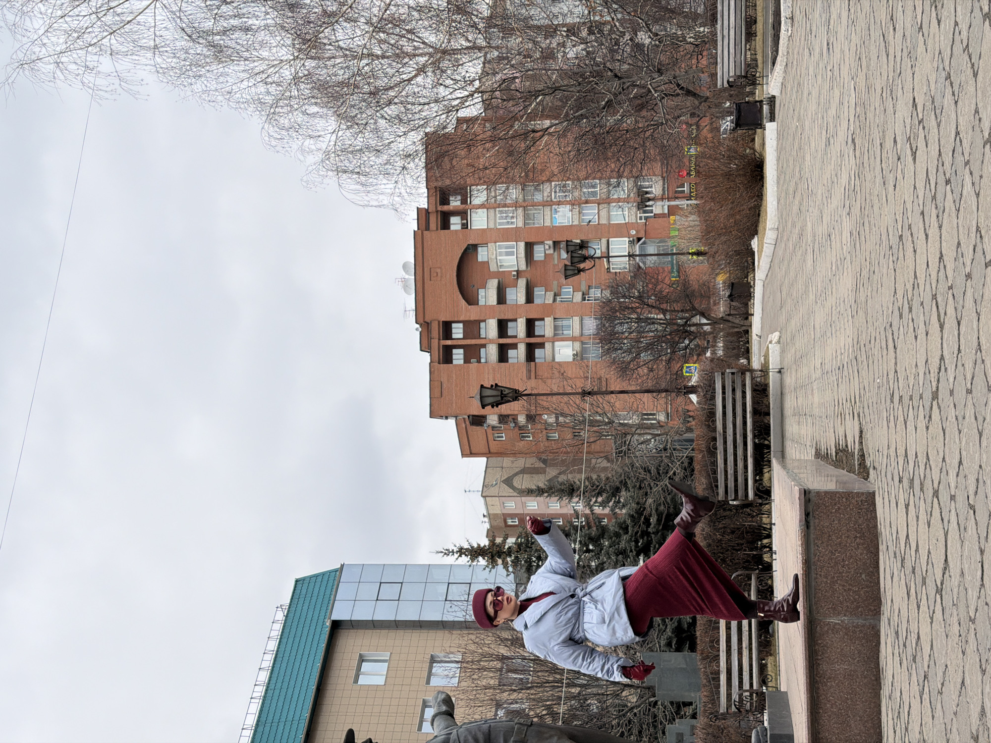 A street scene in Krasnoyarsk, Russia features a woman standing on a stone structure, possibly a monument base. She wears a light blue coat, a dark red skirt, matching gloves and a hat. Behind her stand a variety of buildings, including a tall red brick building and a tan building with a green roof. The ground is paved with a diamond pattern and trees with bare branches are seen throughout. The sky is overcast. A street light is visible beside the red brick building. In the foreground is a dark statue, cropped out from the bottom of the image. A couple of benches are also visible in the background.