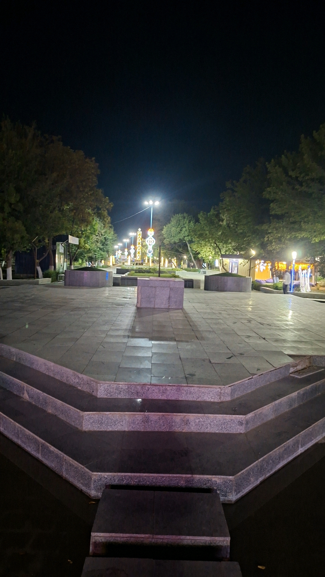 An outdoor nighttime scene at a plaza in Navoiy City, Uzbekistan, features paved ground and tiered steps in the foreground. In the background, trees line a street illuminated by lampposts and decorative lights. Several people are visible walking and standing in the plaza. The overall lighting suggests a public space that is lit for evening use.
