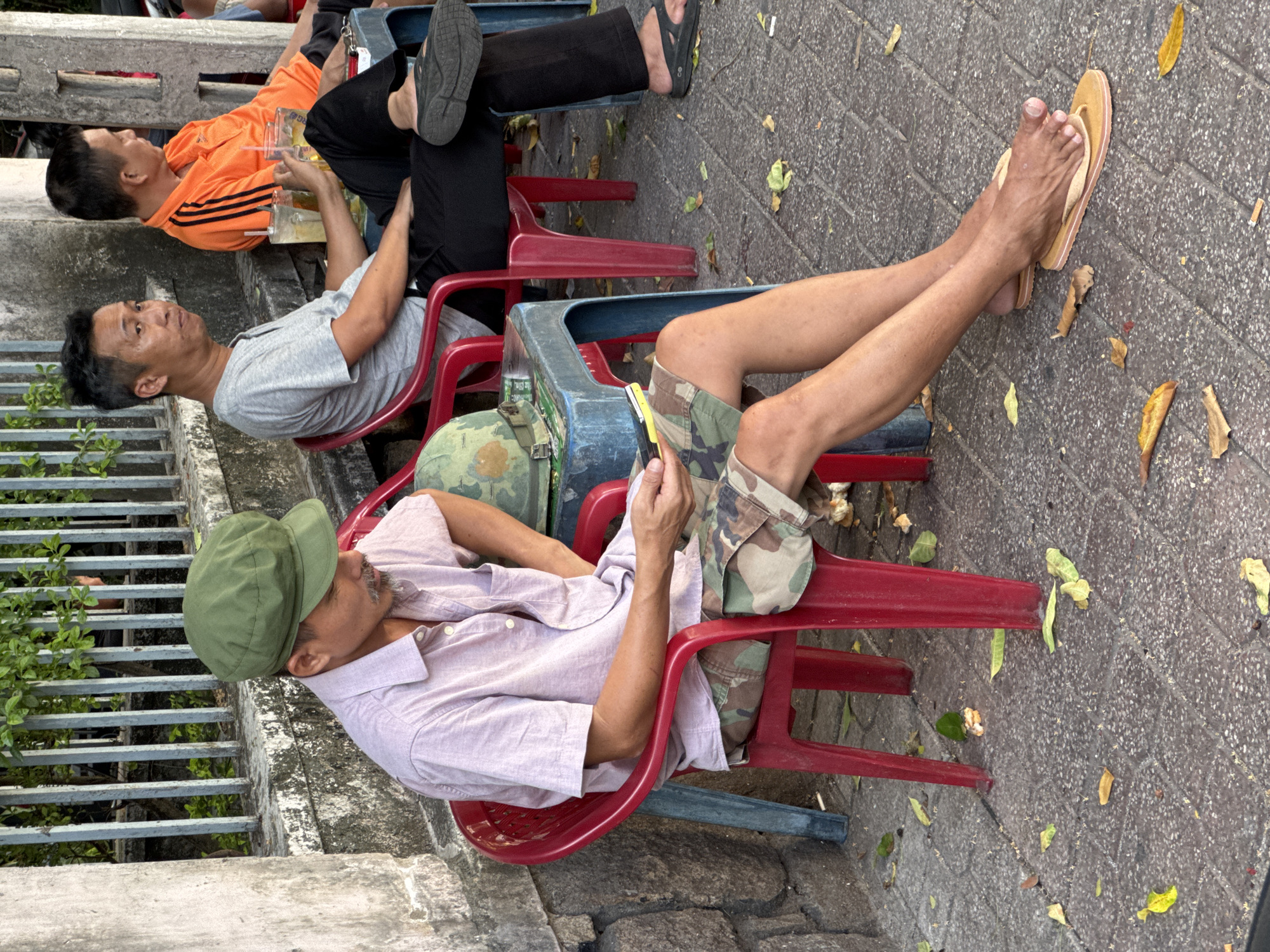 The image depicts a street scene in Nha Trang Ward, Vietnam. Three adult males are seated on plastic chairs, some of them appearing to be at an outdoor cafe or waiting area.
The leftmost individual, wearing a light-colored collared shirt and camouflage shorts, is looking down at a mobile phone he holds in his hand. He also wears a green cap and appears to be using a stylus or similar object with his phone. A military helmet rests on the blue plastic table beside him. His bare feet are resting on flip-flops on the ground.
To his right, another man in a grey t-shirt and black pants is seated, looking towards the viewer with a slight expression. He holds a glass with a beverage and ice in his hand.
Further to the right, a third man is visible, wearing an orange t-shirt with black stripes and black pants. He is looking away from the camera.
The environment appears to be a sidewalk with a paved surface, scattered with fallen leaves. A metal fence and a concrete wall are visible in the background, with greenery peeking through the fence. The overall impression is a casual, everyday scene in a Vietnamese urban setting.