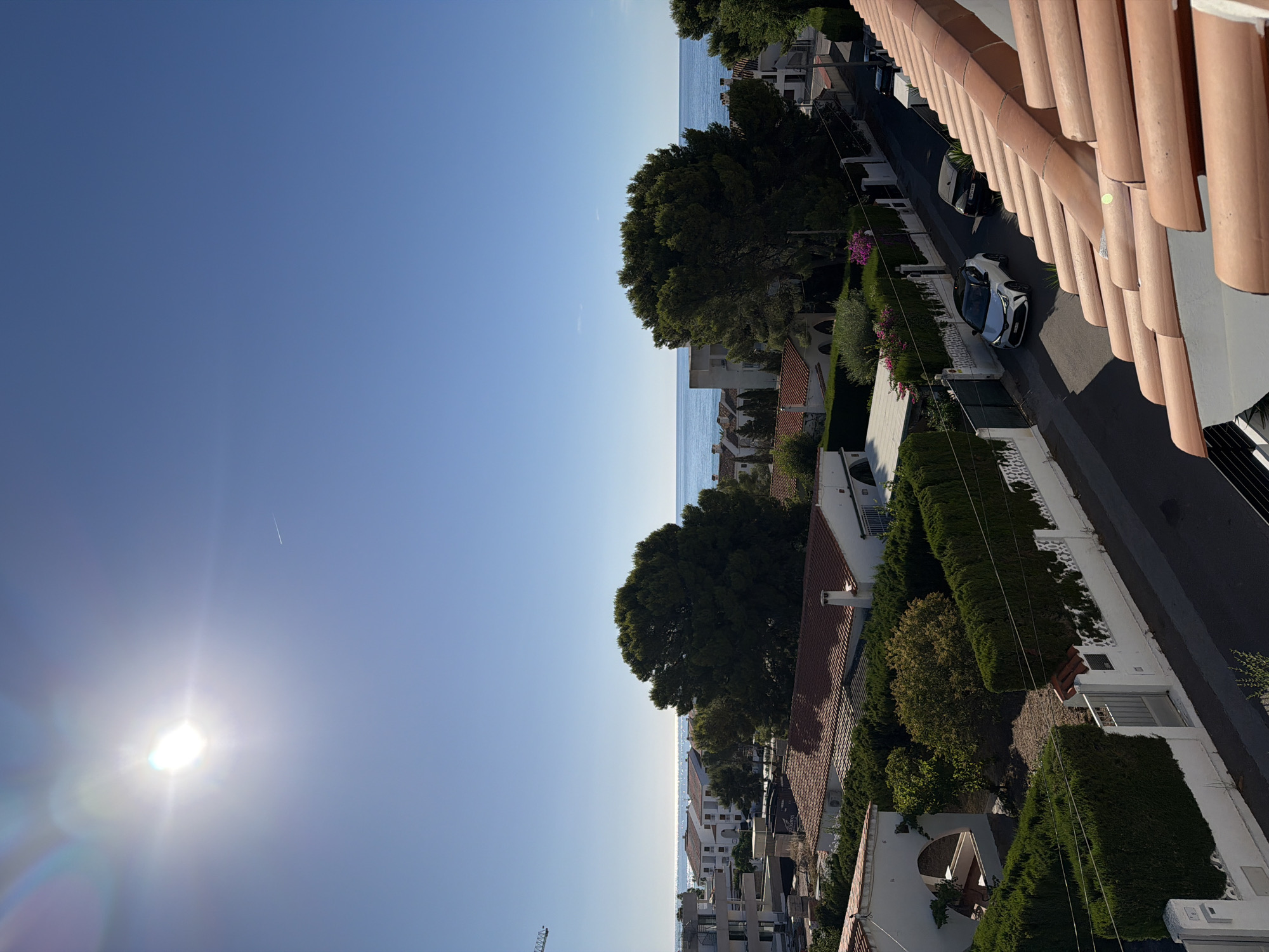The image captures a bright, sunny day in Alcalà de Xivert, Spain, likely from an elevated vantage point, possibly a rooftop or balcony. The foreground is dominated by the terracotta tiles of a roof, sloping downwards towards the right. Beyond the roof, a street lined with white-walled houses and manicured green hedges is visible. Two cars are parked on the street: a white crossover vehicle and a dark-colored SUV. Further in the distance, a line of buildings, some with tiled roofs, are situated before the shimmering blue Mediterranean Sea, which stretches to the horizon under a clear, bright blue sky. The sun is prominently visible in the upper left quadrant of the sky, casting a strong glare and lens flare. A faint contrail from an aircraft is visible in the sky. The overall scene depicts a residential area by the coast on a clear, warm day.