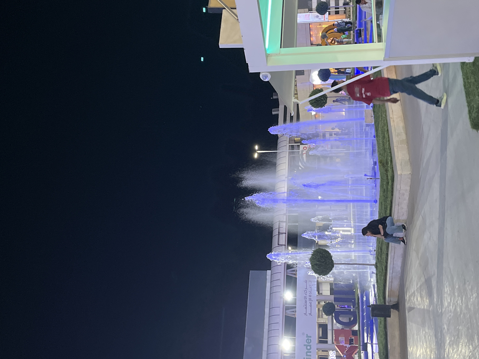 An outdoor fountain plaza is captured at night in Al-Ghizlaniya, Syria. The fountain, illuminated with vibrant blue lights, is the central focus, with water jets reaching high into the dark sky. In the foreground, a man in a red shirt and blue jeans walks away from the camera. To the left of the fountain, a woman with dark hair sits on a stone ledge, looking to the side. Behind the fountain, a building facade is visible with Arabic text, indicating a commercial or business area. The overall scene depicts a lively public space at night, with people enjoying the illuminated fountain.