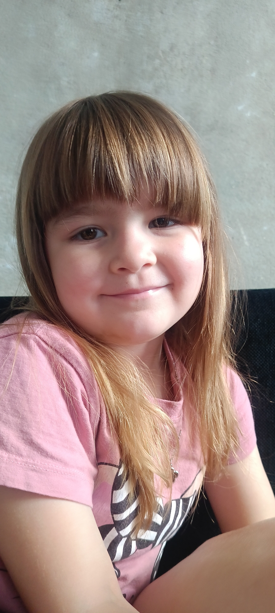 A young girl with shoulder-length, light brown hair and bangs smiles at the camera. She is wearing a pink t-shirt with a graphic print on the front. The background is a textured grey wall and a dark surface, possibly a couch. The lighting suggests it is daytime. The girl's expression is happy and relaxed. The image appears to be a close-up, head-and-shoulders shot. The location context provided is "Хохлы, Russia." There is no visible text in the image.