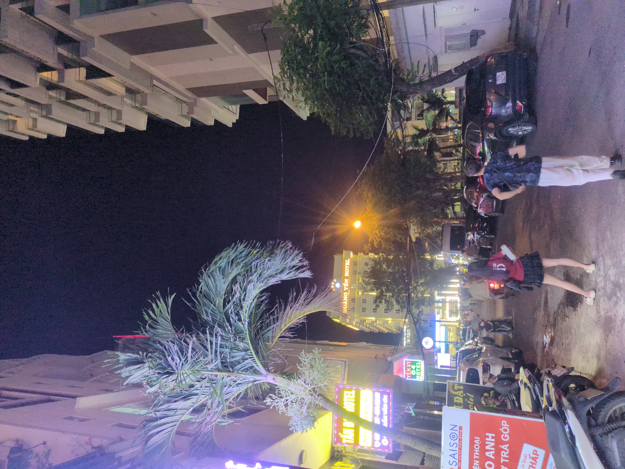 The image depicts a nighttime street scene within Quy Nhon Ward, Vietnam. The scene is illuminated by numerous artificial light sources, including streetlights, building lights, and glowing neon signs.
The street, appearing to be asphalt, extends into the distance, with multi-story buildings lining both sides. On the left, a building hosts "TÂN ĐÔNG HOTEL" and "YẾN VY 04 HOTEL" signs, prominently displaying Vietnamese text. A large red and white sign for "SAISON" and "ĐIỆN THOẠI TRẢ GÓP" (phone installment plan) is visible in the left foreground. Further down the street, the "HOÀNG YẾN HOTEL" is prominently lit. The building on the right features multiple balconies or recessed architectural elements.
Vegetation includes a large palm tree in the left-mid ground and several other trees with dense foliage along the street and adjacent to buildings. Overhead power lines and cables crisscross the upper mid-ground.
Several vehicles are present. In the left foreground, multiple motorcycles are parked. A silver SUV/crossover and a dark-colored SUV/crossover (license plate 77A-042.35) are parked in the mid-ground. Additional motorcycles and a small van are visible further down the street.
Two individuals are centrally located in the mid-ground, walking away from the camera. The person on the left is female, wearing a dark red short-sleeved top with a white number '5' on the back, a black pleated mini-skirt, and light-colored flat shoes. She holds a white object in her left hand. The person on the right is male, wearing a dark patterned short-sleeved shirt, light-colored loose trousers, dark shoes, and a dark bag slung across his body. In the left-mid background, another individual wearing a cap and mask is partially visible near a parked car. Other pedestrians and individuals on motorcycles are visible further down the street.