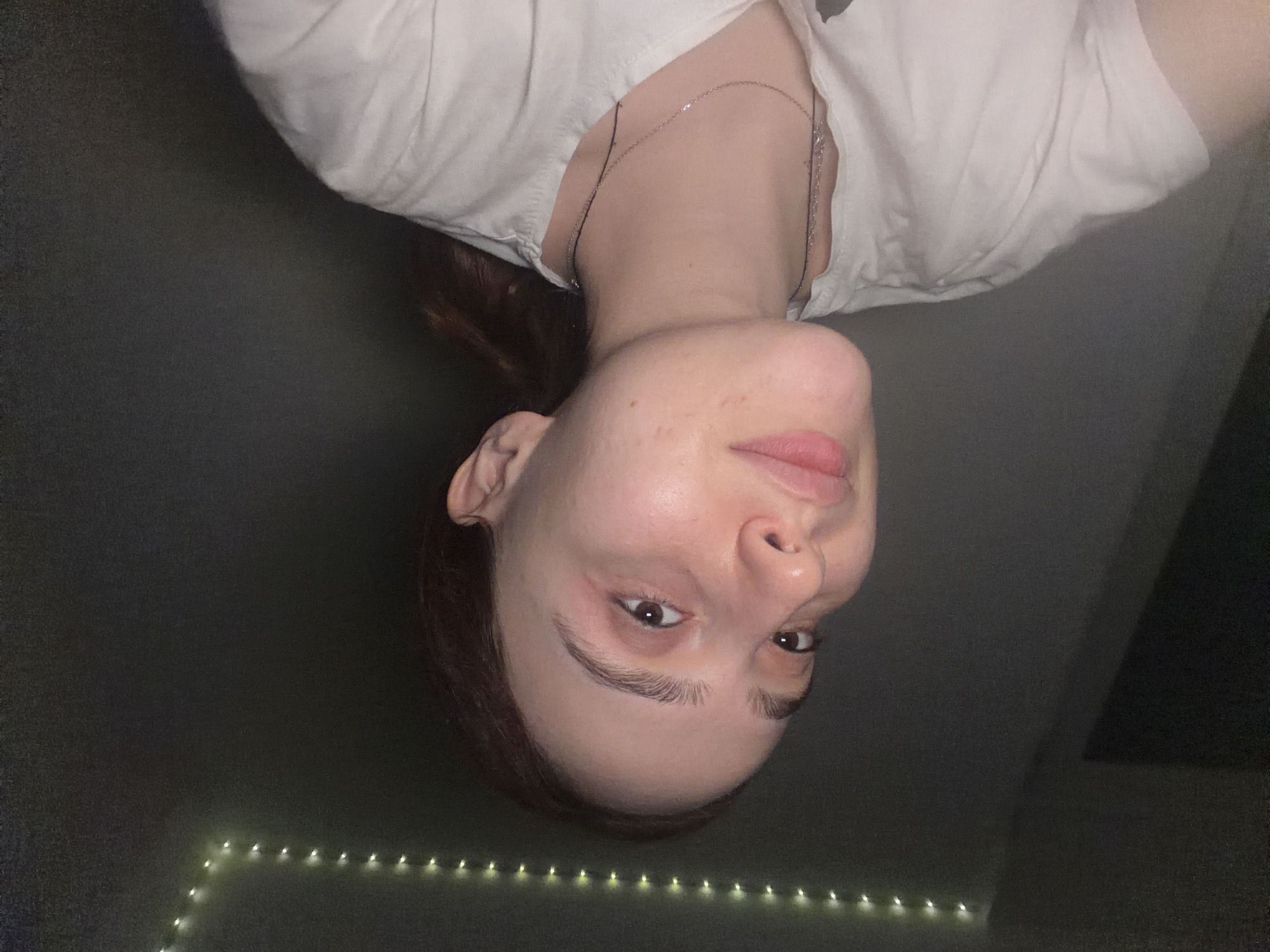 A close-up portrait of a single female individual, appearing to be in her late teens or early twenties, is captured indoors while posing for the photograph. She possesses light skin, brown hair styled away from her face, brown eyes, and light brown eyebrows, exhibiting a subtle upward gaze and a slight smile. She is attired in a white short-sleeved t-shirt and accessorized with two thin necklaces, one silver-colored chain and one dark string. The background is a dark, featureless wall, with a string of illuminated warm white lights running horizontally along its upper section. The ambient lighting is dim. No visual cues within the image indicate its alleged location in Strasbourg, France.