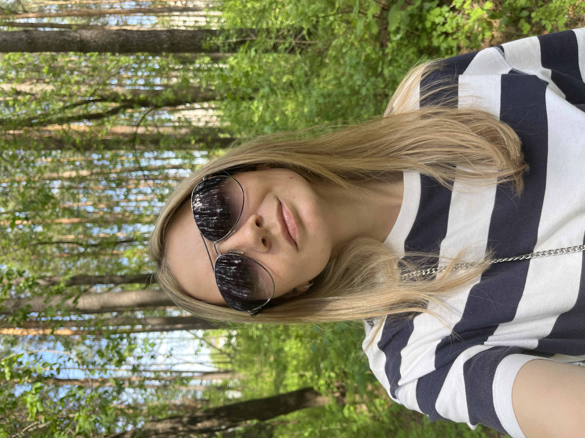 A young woman with blonde hair and sunglasses is taking a selfie outdoors. She is wearing a black and white striped shirt. The background consists of trees with green leaves, suggesting a forest or park setting. The location is likely Chaikovsky, Russia.