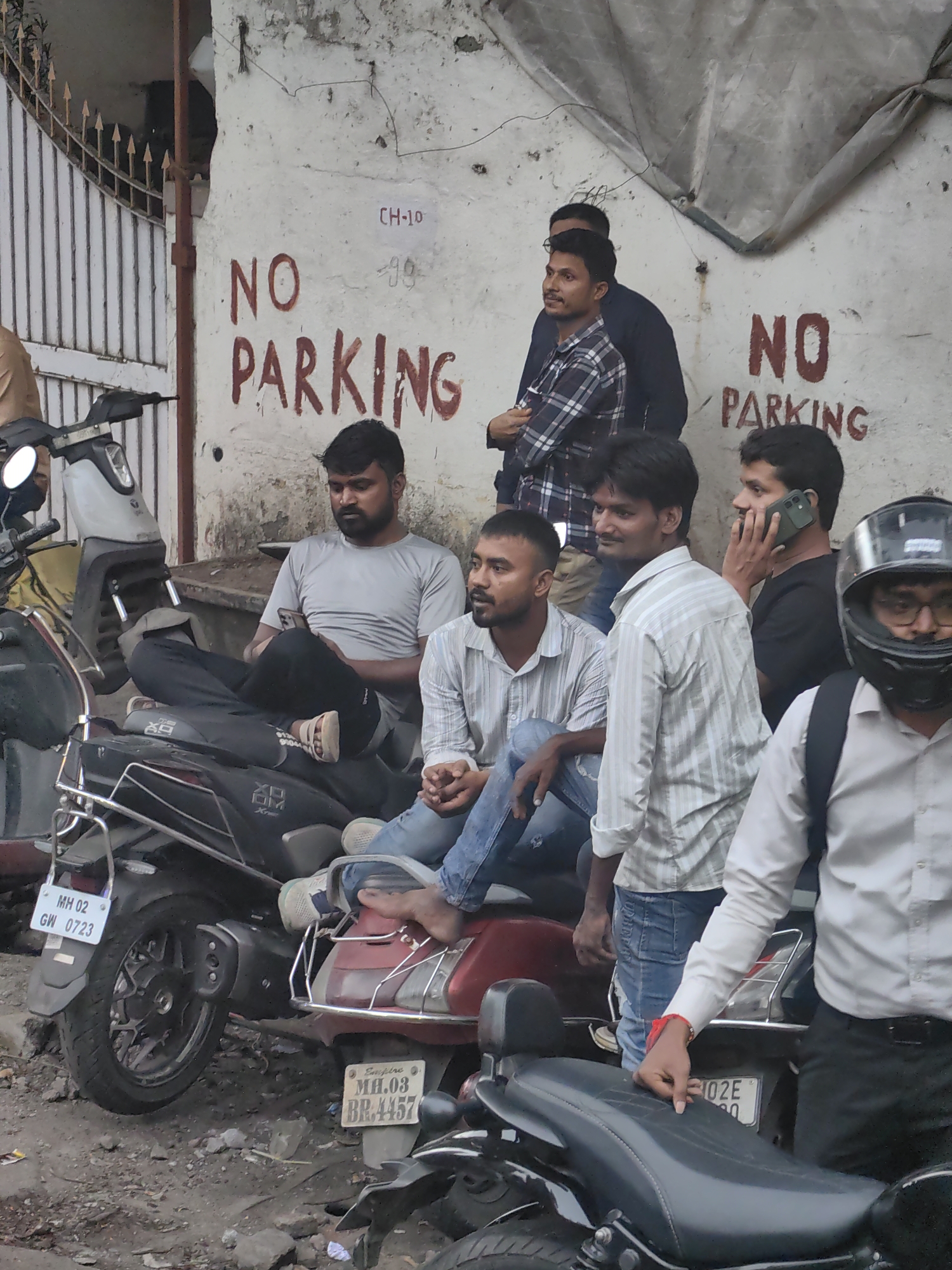 The image depicts a group of men gathered around motorcycles outside a building with a white, weathered wall. Red spray paint on the wall reads "NO PARKING." Some men are seated on the motorcycles, while others are standing. One man is on his phone, and another is wearing a motorcycle helmet. The ground is rough and uneven, with debris scattered around. The scene appears to be in a busy urban environment, possibly in Mumbai, India, given the license plates on the motorcycles which start with "MH," the abbreviation for Maharashtra state. The overall atmosphere is casual and relaxed, with the men appearing to be socializing or waiting.