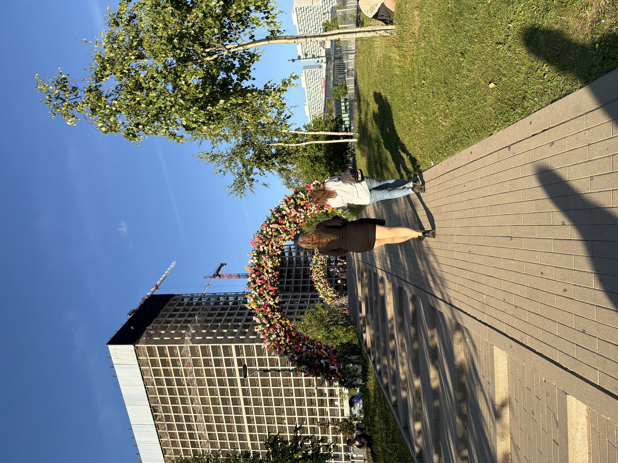In an outdoor urban environment under a clear blue sky, two women walk away from the camera on a paved walkway. The woman in front wears a short black dress and black heels. The woman behind her wears a white t-shirt, blue jeans, and a backpack. To their left, a wide set of concrete stairs ascends, bordered by a grassy slope. Ahead of them, a large floral archway, made of numerous colorful flowers, spans across the walkway, leading towards a bridge. Behind the archway, a large, multi-story building under construction dominates the background, with cranes visible. To the right of the walkway, a grassy area is interspersed with trees. In the far background, several modern apartment buildings are visible. The sun casts long shadows from the trees and people onto the walkway and grass.