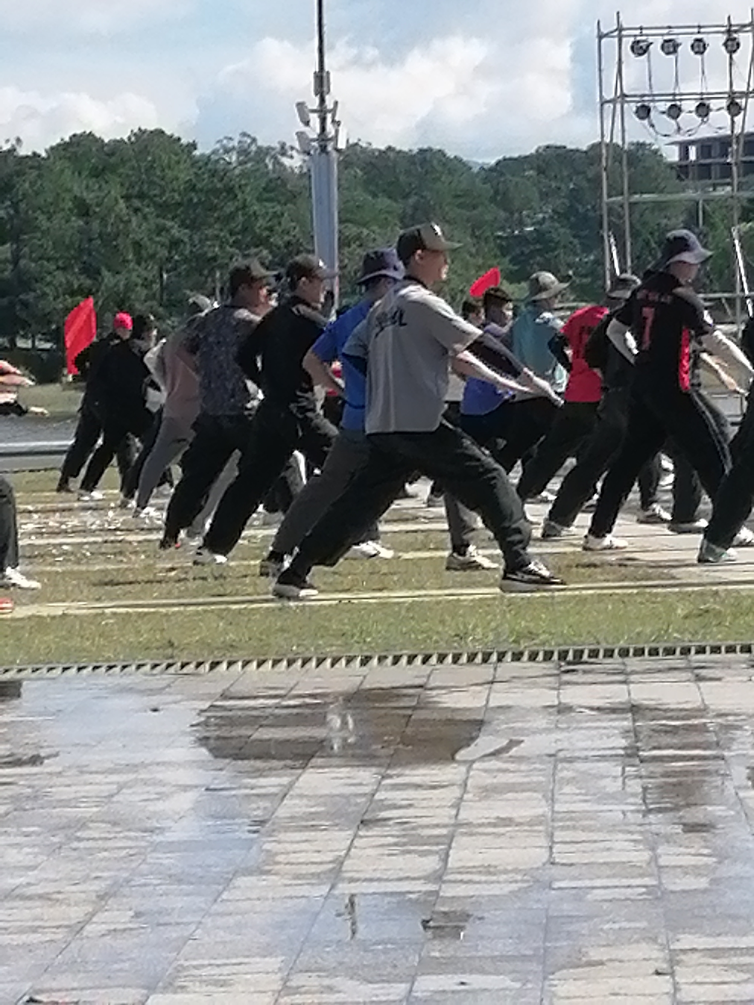 The image depicts a group of people engaged in an outdoor activity, possibly a synchronized exercise or performance, on a paved area with grass in the background. The participants are dressed in casual athletic wear, primarily dark pants and shirts, with some wearing hats. Their movements suggest a coordinated effort. In the background, trees, a tall mast with antennas, and a metal scaffolding structure with lights are visible, indicating an urban or park-like setting. A red flag is also present, adding a splash of color to the scene. The paved ground shows reflections, suggesting it is wet from rain or cleaning. The scene is set during daylight hours, with a cloudy sky overhead. The activity appears to be taking place in a public space, potentially a plaza or park. This scene does not appear to be in Da Lat, Vietnam.