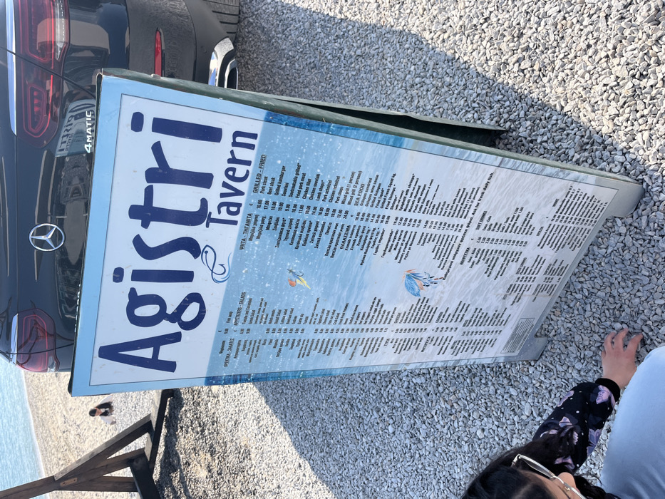 The image shows a sign for "Agistri Tavern" in Limenaria, Greece (coordinates: 40.626, 24.569). The sign is white with blue lettering and displays a menu of food and drinks. The sign is located on a gravel pathway. A woman is crouched on the ground in the lower left corner of the image. A black car is partially visible in the upper right corner, with a "4MATIC" logo on the rear bumper. The sign is slightly tilted and partially obscured by the car. The sign is lit by sunlight, and the gravel path is visible in the foreground and background. The menu lists a wide variety of dishes, including appetizers, salads, pita, grilled, fried, and seafood. There is also a section for soft drinks and wine. The environment is likely a restaurant or tavern area. The woman is looking down at the ground and appears to be reading the menu.