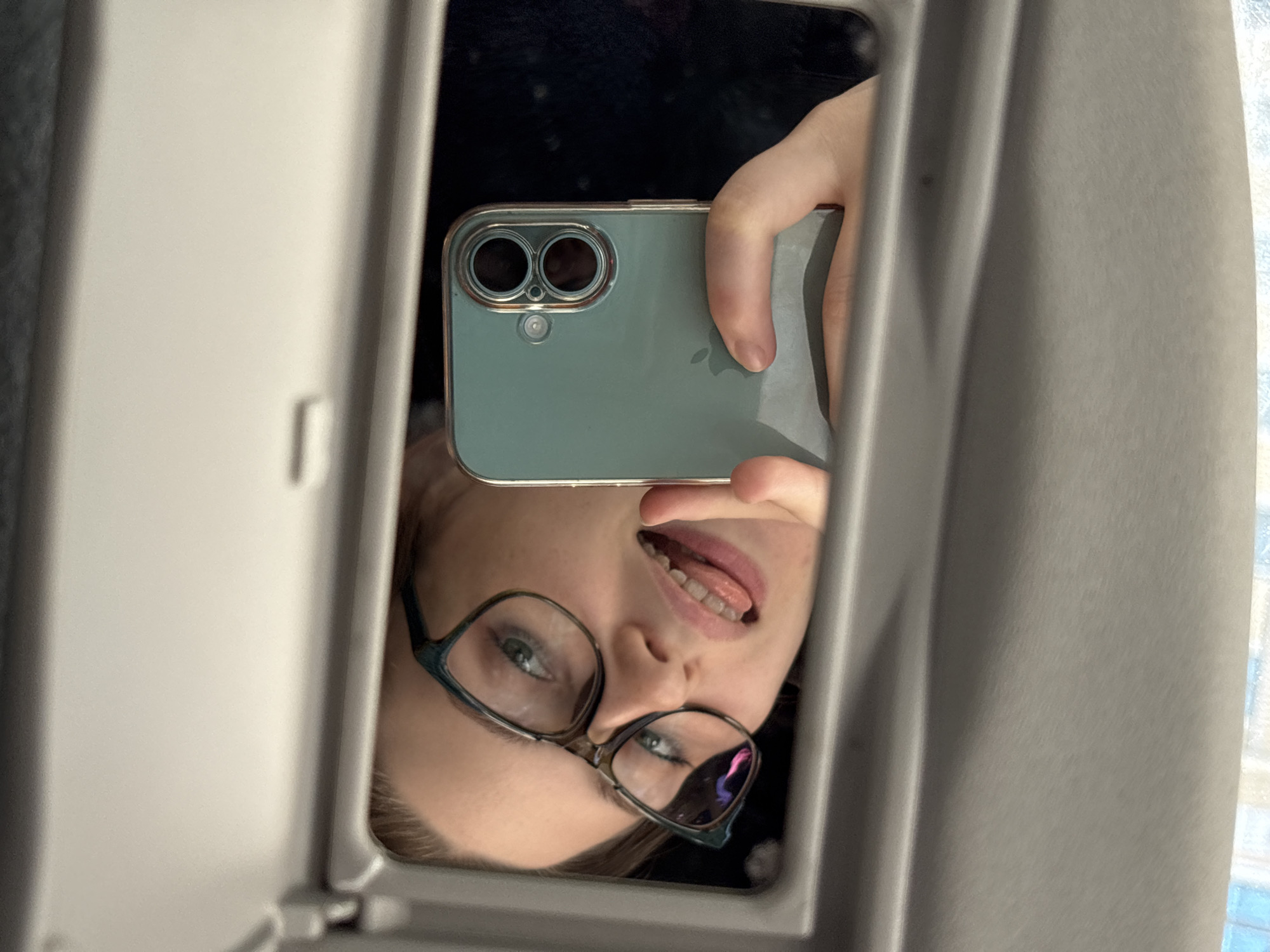 The image shows a mirror selfie taken by a young person wearing glasses. They are holding a smartphone with a light blue case, and the phone's camera is pointed towards the mirror. The person is sticking their tongue out and appears to be making a playful expression. The reflection in the mirror captures their face, hair, and the smartphone they are using. The background is dark and blurry, suggesting an indoor setting, possibly a car, given the visible interior surfaces which are gray and textured. There is no visible text in the image. The lighting suggests it might be daytime, but the weather and specific location cues within Saint Petersburg are not discernible from the image itself.
