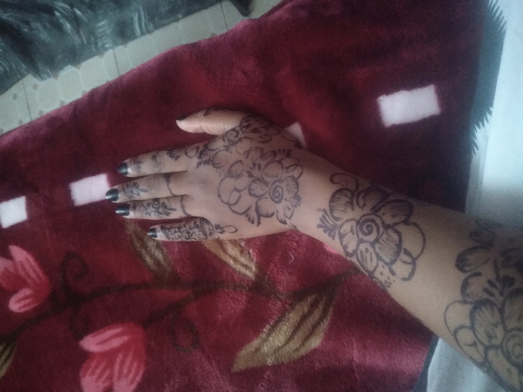A person's arm and hand are adorned with henna tattoos, depicting floral designs. The fingernails are painted black. The arm is resting on a maroon blanket with white patches and floral patterns. The location is unknown.