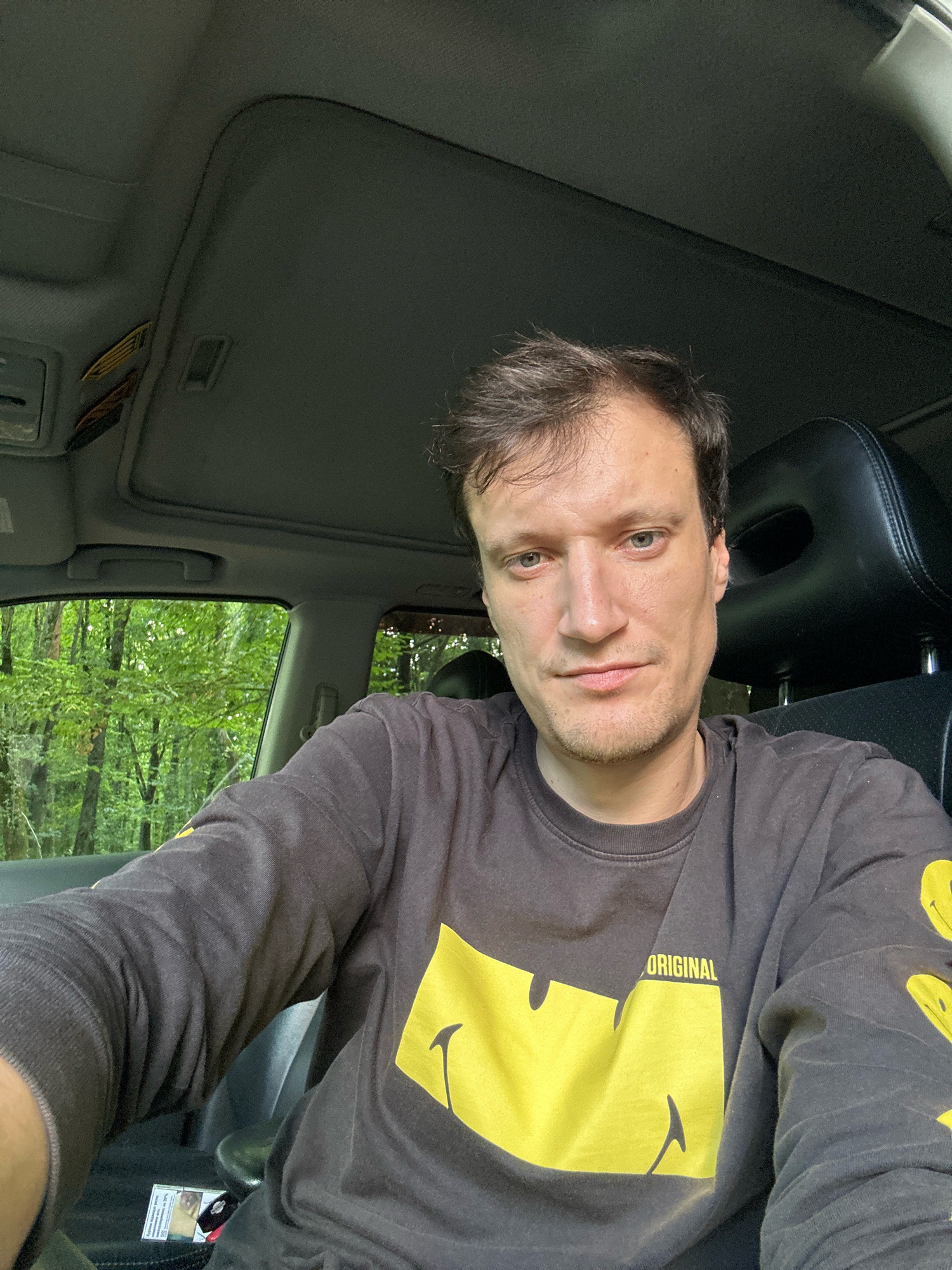 A man is seated in the driver's seat of a car, with a selfie taken from a low angle. The car interior is visible, including a sunshade on the ceiling and the black headrest of a seat. Through the open window, a dense forest with green foliage can be seen. The man is wearing a dark grey sweatshirt with a large yellow smiley face design and the word "ORIGINAL" printed on it. A pack of cigarettes is visible on the console in front of him. The scene appears to be set in a natural environment, likely a forest road, possibly near Shatava, Ukraine, given the context.