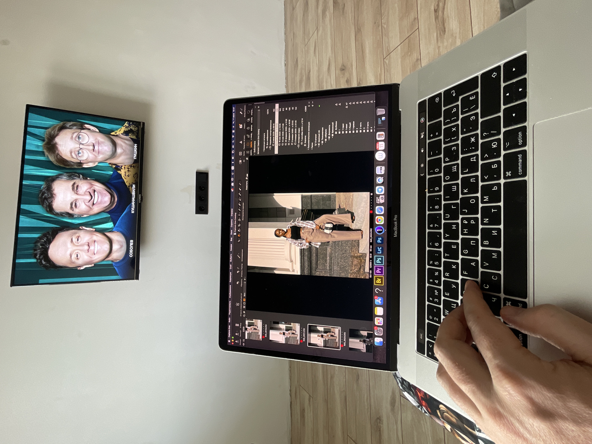 The image shows a person's hand on the keyboard of a MacBook Pro, with the laptop displaying photo editing software. A TV screen mounted on a wall above the laptop shows three men's faces. The setting appears to be indoors, with a wooden floor visible. The TV screen displays the names "СОБОЛЕВ", "КУШАНАШВИЛИ", and "ПУШКИН" beneath the men's images, suggesting a Russian context. The person's hand is positioned as if typing or interacting with the keyboard. The overall scene is a snapshot of someone working on their laptop with entertainment playing on the TV. There is no indication of the city of Kazan, Russia, or any specific location within it.