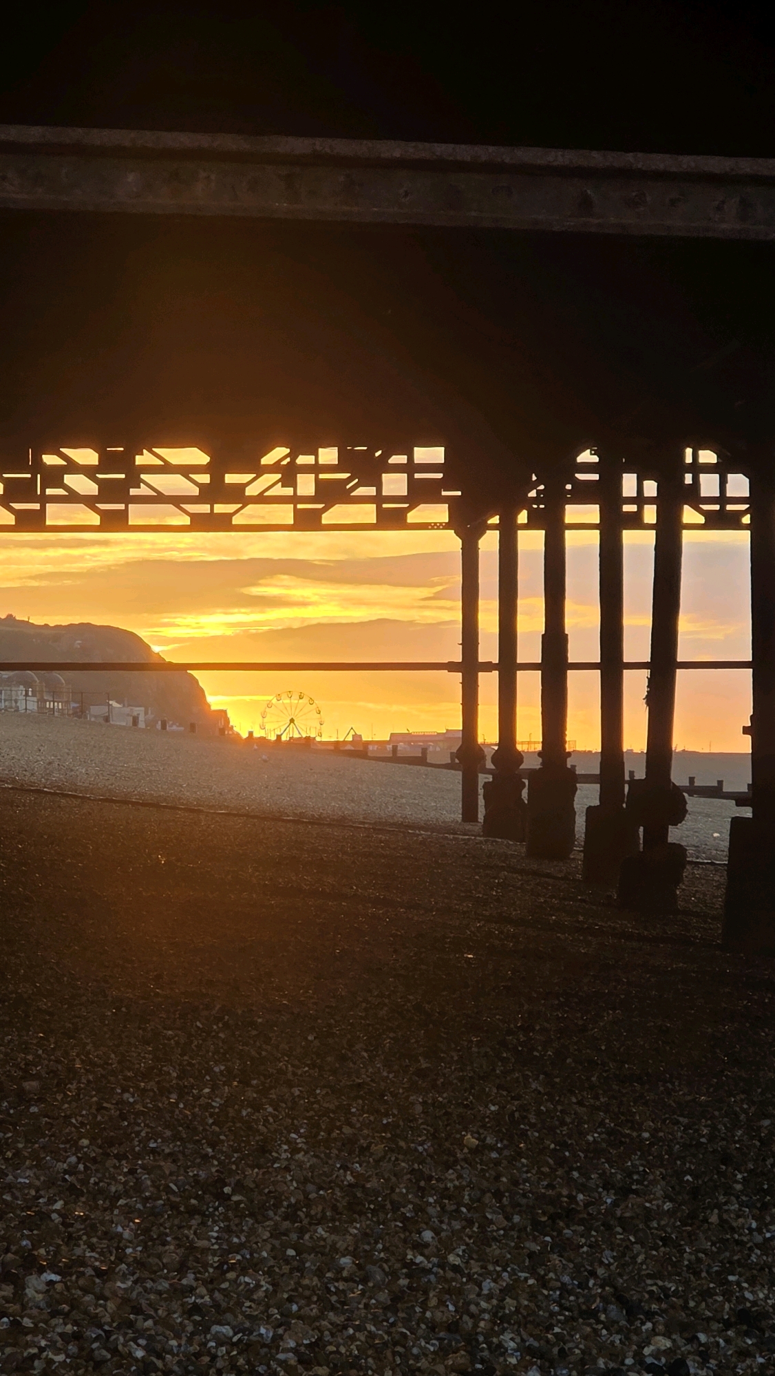The image shows a view of the beach and coastline in St Leonards-on-Sea, United Kingdom, at sunset. The perspective is from underneath a pier structure, with its dark supports and lattice ceiling visible in the foreground and upper frame.
The sunset casts a vibrant orange and yellow glow across the sky, with clouds diffused by the light. In the middle ground, a pebble beach stretches towards the sea. A Ferris wheel is visible on the horizon, along with buildings and structures along the promenade. A gentle slope of land, covered in buildings and greenery, rises on the left side of the image. The sea itself is not prominently visible, but its presence is implied by the beach and the distant coastline. The lighting is low due to the sunset, creating strong shadows and silhouettes from the pier structure. There are no people or animals clearly depicted in the scene. The overall mood is serene and captures the end of the day by the coast.