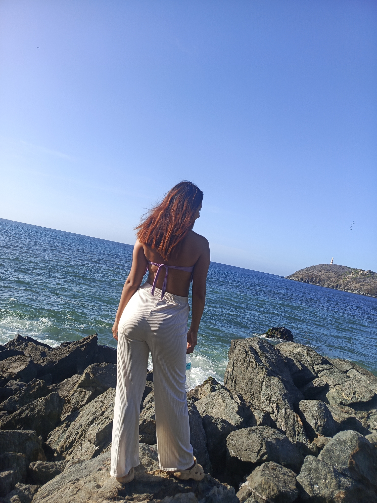 A woman with long, auburn hair stands on a rocky shore, facing away from the camera and looking out at the ocean. She is wearing a purple bikini top and loose, cream-colored pants. The water is blue and appears somewhat choppy, with whitecaps near the shore. In the distance, a lighthouse is visible on a small, rocky island. The sky is clear and blue. The location is identified as Pedro González, Venezuela.