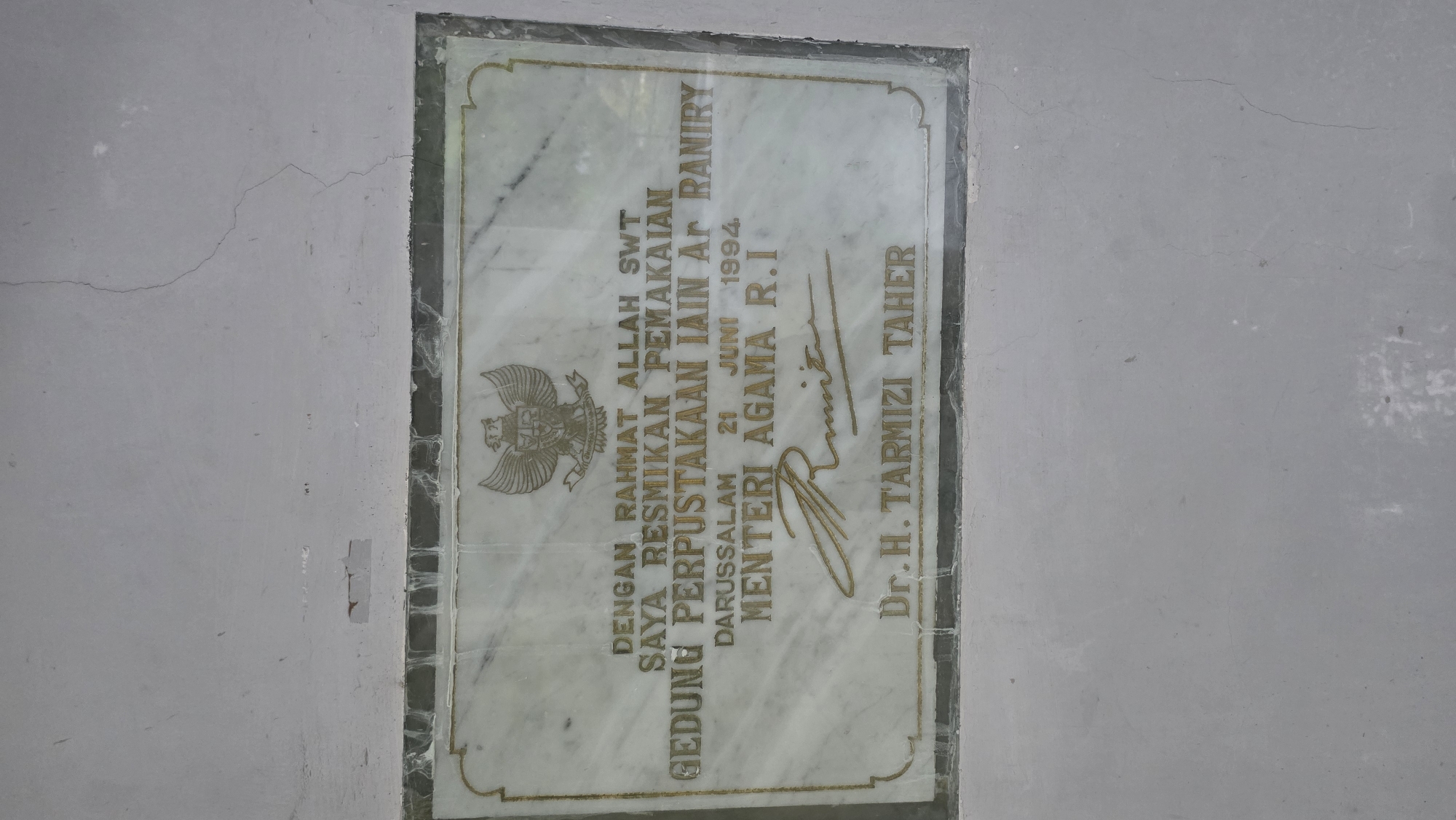 The image shows a marble plaque mounted on a wall. The plaque commemorates the inauguration of a library building at IAIN Ar-Raniry on June 21, 1994, in Darussalam. The text indicates that the inauguration was done with the grace of Allah SWT and it was officiated by Dr. H. Tarmizi Taher, the Minister of Religious Affairs of the Republic of Indonesia. The plaque features an emblem similar to Garuda Pancasila. The wall appears to be painted light gray and has a crack running across its surface.