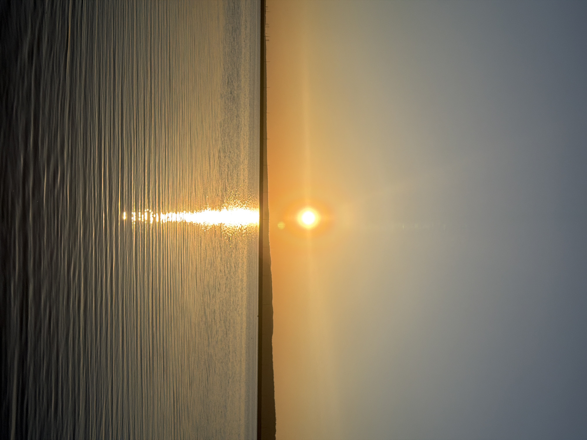 The image shows a sunset over the water. The water surface is rippled, reflecting the sunlight. The sky is a gradient of orange and blue. The location is likely near the city of Chaikovsky, Russia, as indicated in the prompt. The image portrays a serene and natural scene.