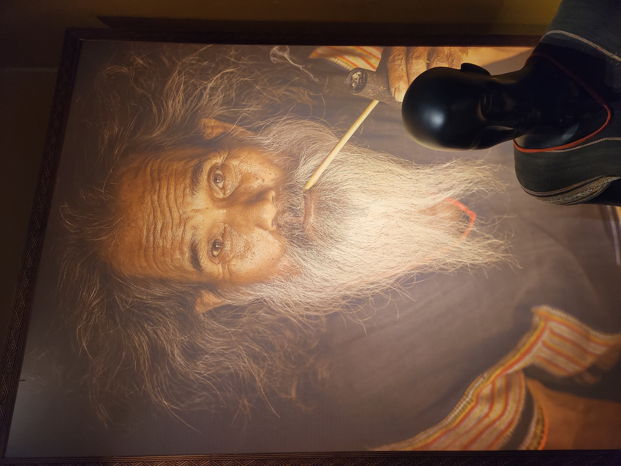 A framed portrait of an elderly man with a long white beard and wispy hair is displayed on a wall. He is smoking a pipe, with smoke gently rising from it. The man is wearing a traditional garment with striped patterns. In the foreground, a black mannequin is positioned in front of the portrait, obscuring part of the man's lower face and chest. The mannequin is wearing a similar garment with striped detailing around the neckline. The background appears to be a wall or display area within a museum or gallery in Hội An Ward, Vietnam.