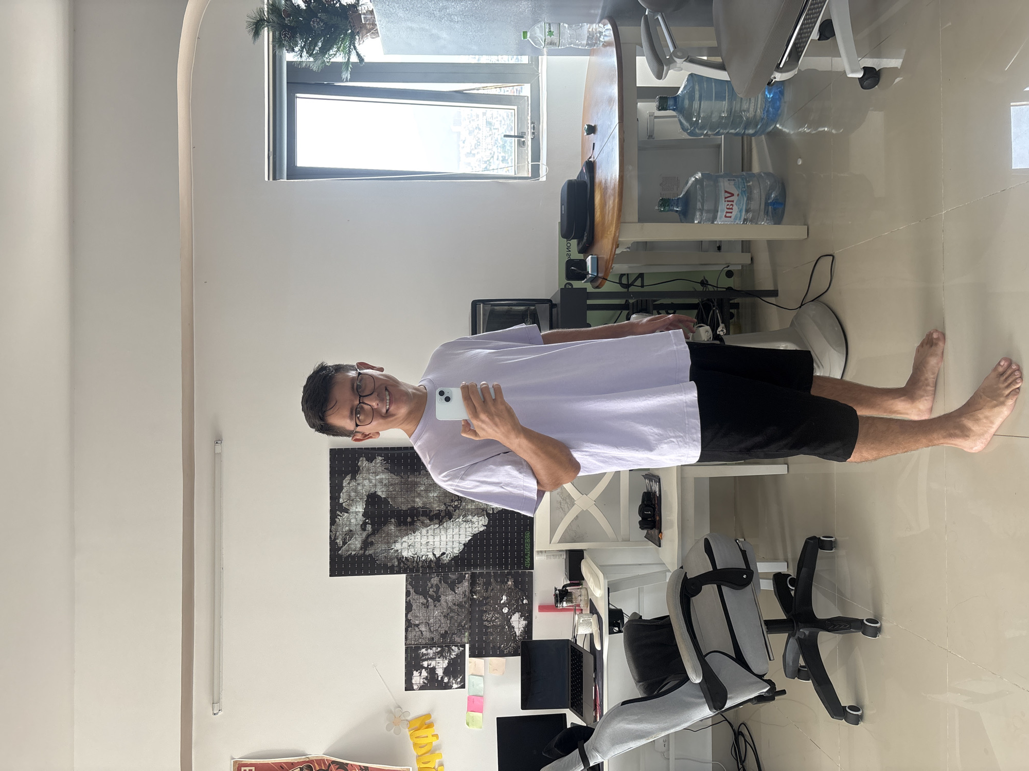 A young man with glasses stands in a well-lit room, holding a smartphone in his right hand and taking a mirror selfie. He is wearing a light purple t-shirt and black shorts, and is barefoot. The room appears to be a home office or living space, with a desk equipped with two monitors, a laptop, and a pink cylindrical object. Behind the desk, there are several framed black and white posters on the wall, along with some colorful sticky notes. To the right of the desk, a white chair with a patterned seat is visible. Further to the right, a round wooden table is set up, with two large water jugs beneath it. A window offering a view of a city skyline can be seen in the background, and a small decorative Christmas tree is positioned near the window. The floor is tiled and reflective, and a grey office chair is positioned to the left of the desk.