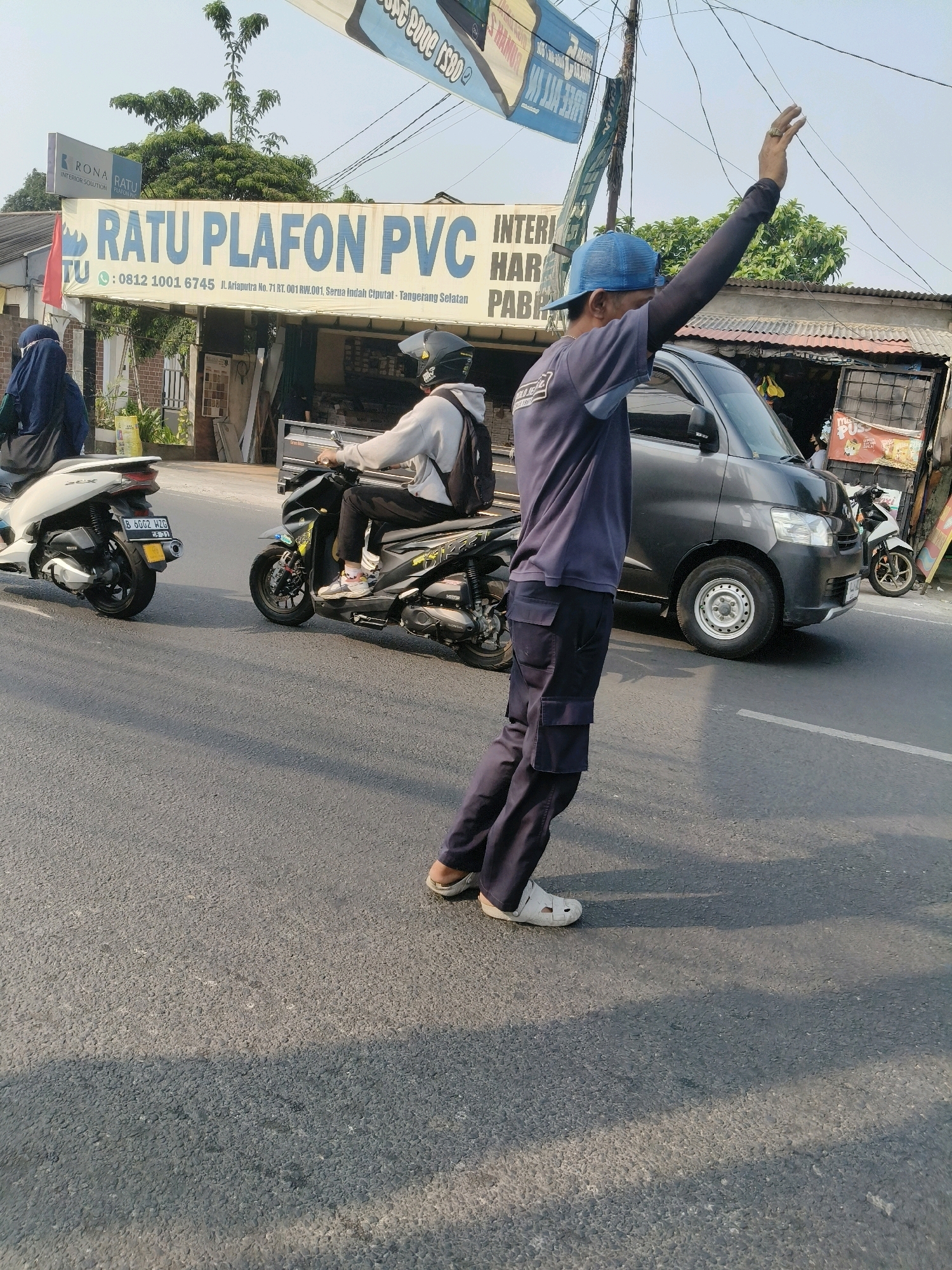 A male traffic director, wearing a blue baseball cap, a dark blue long-sleeved shirt with dark blue cargo pants, and white crocs, stands in a street in South Tangerang, Indonesia. He is gesturing with his right hand raised, possibly directing traffic or greeting someone. In the background, a dark gray minivan is parked. To the left of the minivan, a person on a motorcycle wearing a gray hoodie and a black backpack is visible. A white scooter is parked further to the left. A storefront with a banner that reads "RATU PLAFON PVC" is situated behind the individuals. The scene takes place on a paved road under a clear sky, with trees and buildings lining the street.
