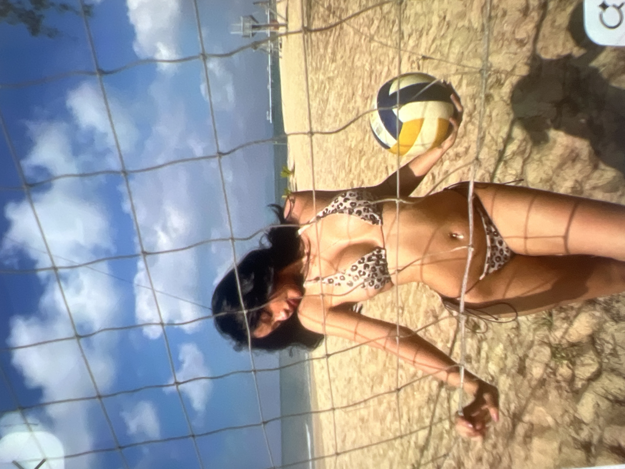 The image shows a woman wearing a leopard-print bikini on a sandy beach. She is holding a volleyball and standing behind a volleyball net. The background shows the ocean and a clear blue sky with clouds. There are no identifiable landmarks that would place this image specifically in Kara-Dıykan, Kyrgyzstan.