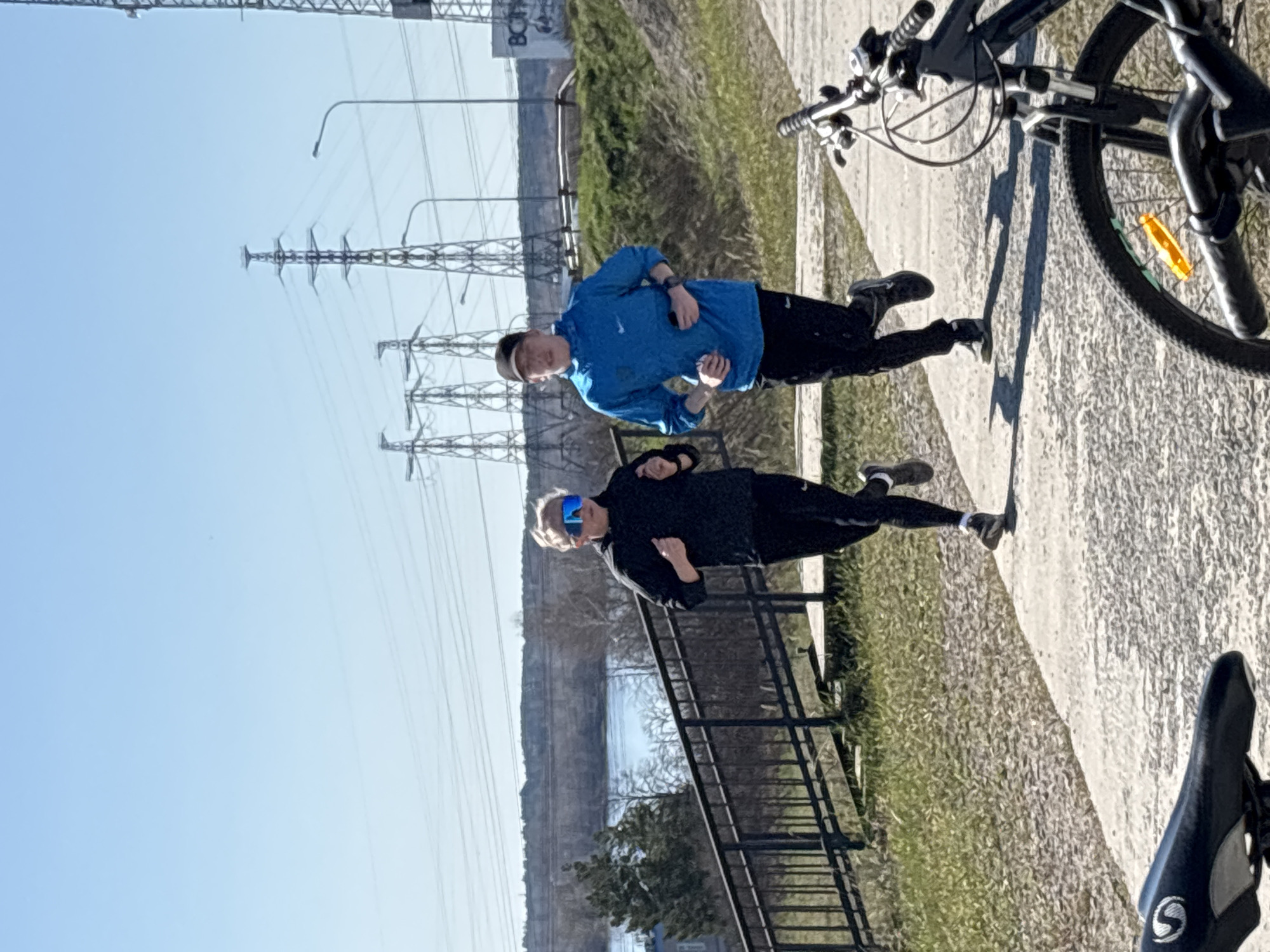 Here's a detailed description of the image:
The image shows two people, likely a man and a woman, standing on a paved path. They appear to be engaged in some sort of physical activity or stretching. The man is wearing a blue long-sleeved shirt and black pants, while the woman is wearing a black tracksuit and blue sunglasses. The photo features an outdoor setting; in the background, there are utility poles and power lines, indicative of an urban or semi-urban landscape. There is a railing along the pathway, behind which are trees and a body of water. A bicycle, with its front wheel partially visible, is present on the right side of the image. The scene is bathed in sunlight, indicating it is daytime. Considering the presence of power lines and a river, it is likely the image was taken in an industrial or residential area in Chaikovsky, Russia.