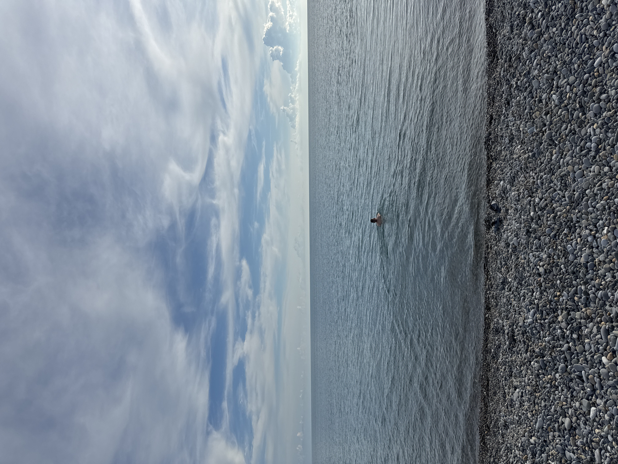 A person is swimming in the calm, rippling waters of the Black Sea, seen from a pebble beach. The sky above is a vast expanse of blue with scattered, wispy white clouds. The horizon is sharply defined where the water meets the sky. The foreground is dominated by the dark, rounded stones of the beach. The scene suggests a peaceful moment by the sea in Sochi, Russia.