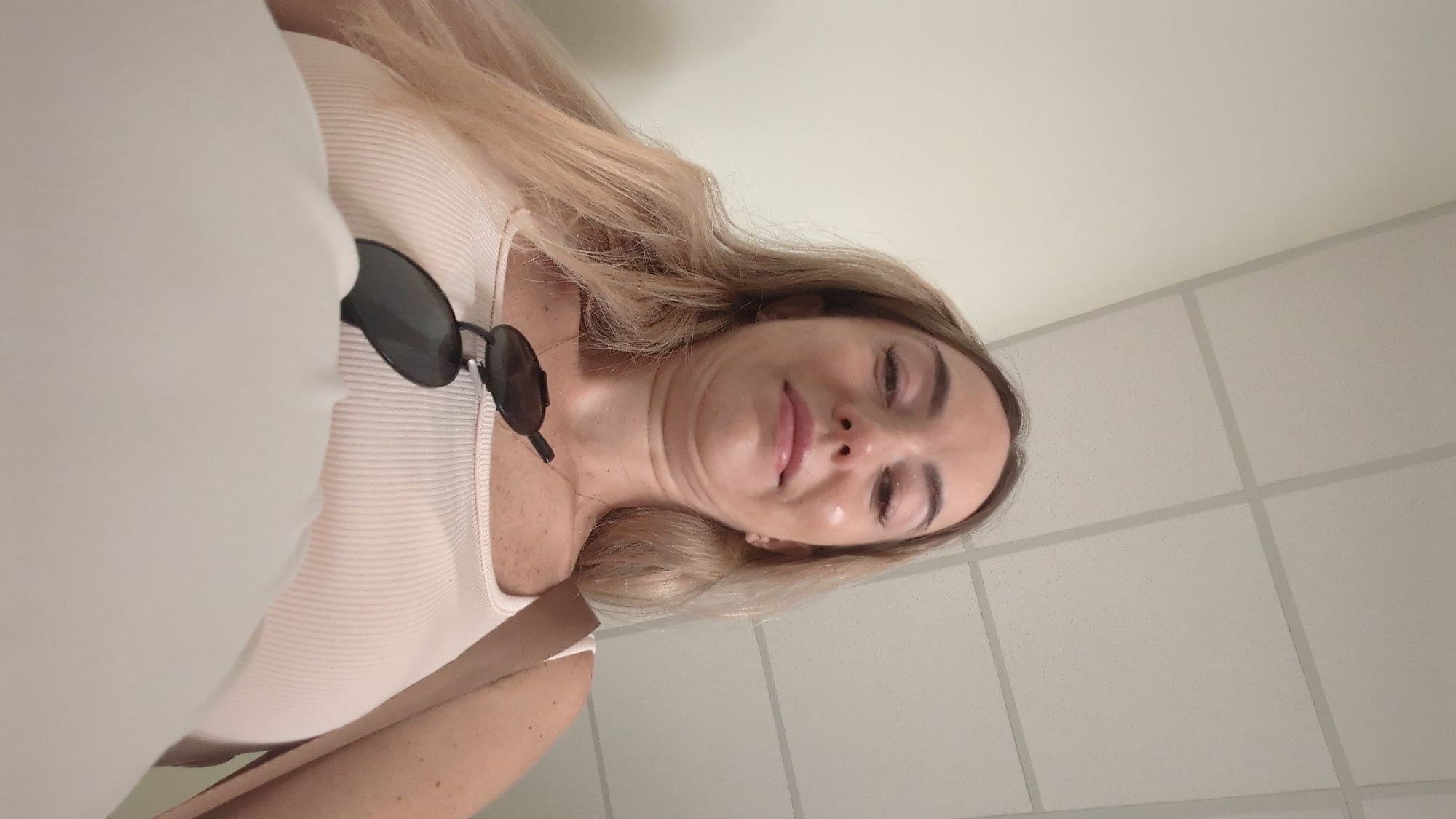 An indoor selfie captures a woman looking down at the camera. She has blonde hair and is wearing a light pink ribbed tank top with sunglasses resting on it. A light brown strap from a bag is visible over her shoulder. The background consists of a plain, light-colored wall and a tiled ceiling with grid lines. There is no indication of the specific location within Chișinău, Moldova.