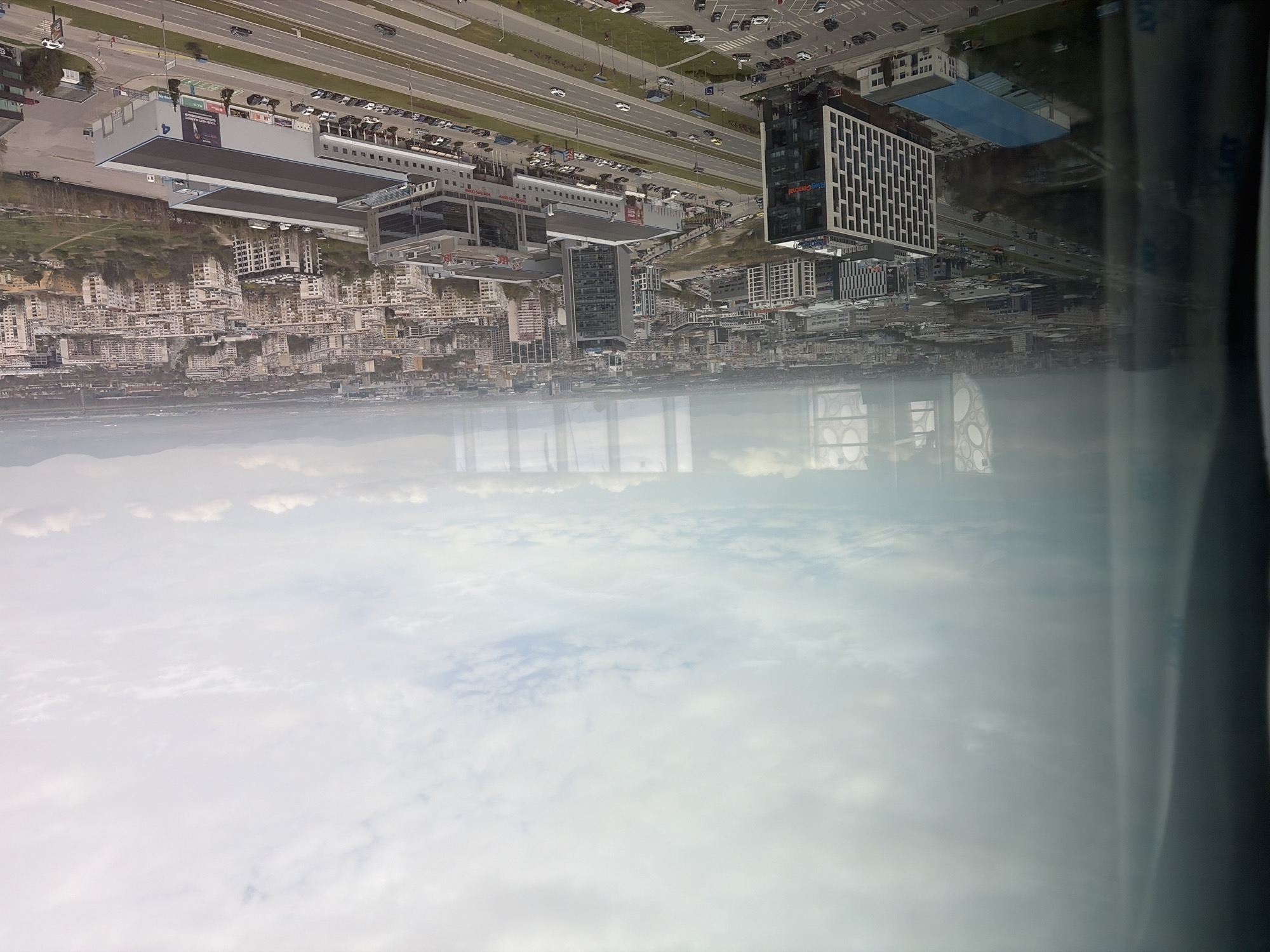 An aerial-like view captures a sprawling cityscape in Sofia, Bulgaria, taken from a high vantage point through a window, as indicated by reflections of internal window frames and the sky in the lower portion of the image.
The scene depicts a vibrant urban environment under an overcast sky with scattered clouds, suggesting daytime. In the midground, a multi-lane highway stretches across the scene, bordered by numerous vehicles parked in adjacent lots. A mix of residential and commercial buildings dominates the landscape.
To the left of the highway, a large, modern building complex with a flat roof and extensive parking areas is prominent. A visible sign on its roof displays the number "4", and another sign features the text "YOUR NEXT TRAVEL". To the right of the highway, a distinctive, multi-story office building stands out with a patterned white facade and clearly visible red text reading "Ring Central". The area around these large structures is densely populated with many apartment buildings, forming a modern urban fabric.
In the far background, distant mountains are visible, partially obscured by atmospheric haze. The reflection on the window in the foreground shows distorted images of the sky and light. The image captures a dynamic urban landscape, showcasing a blend of infrastructure, commerce, and residential areas in Sofia.