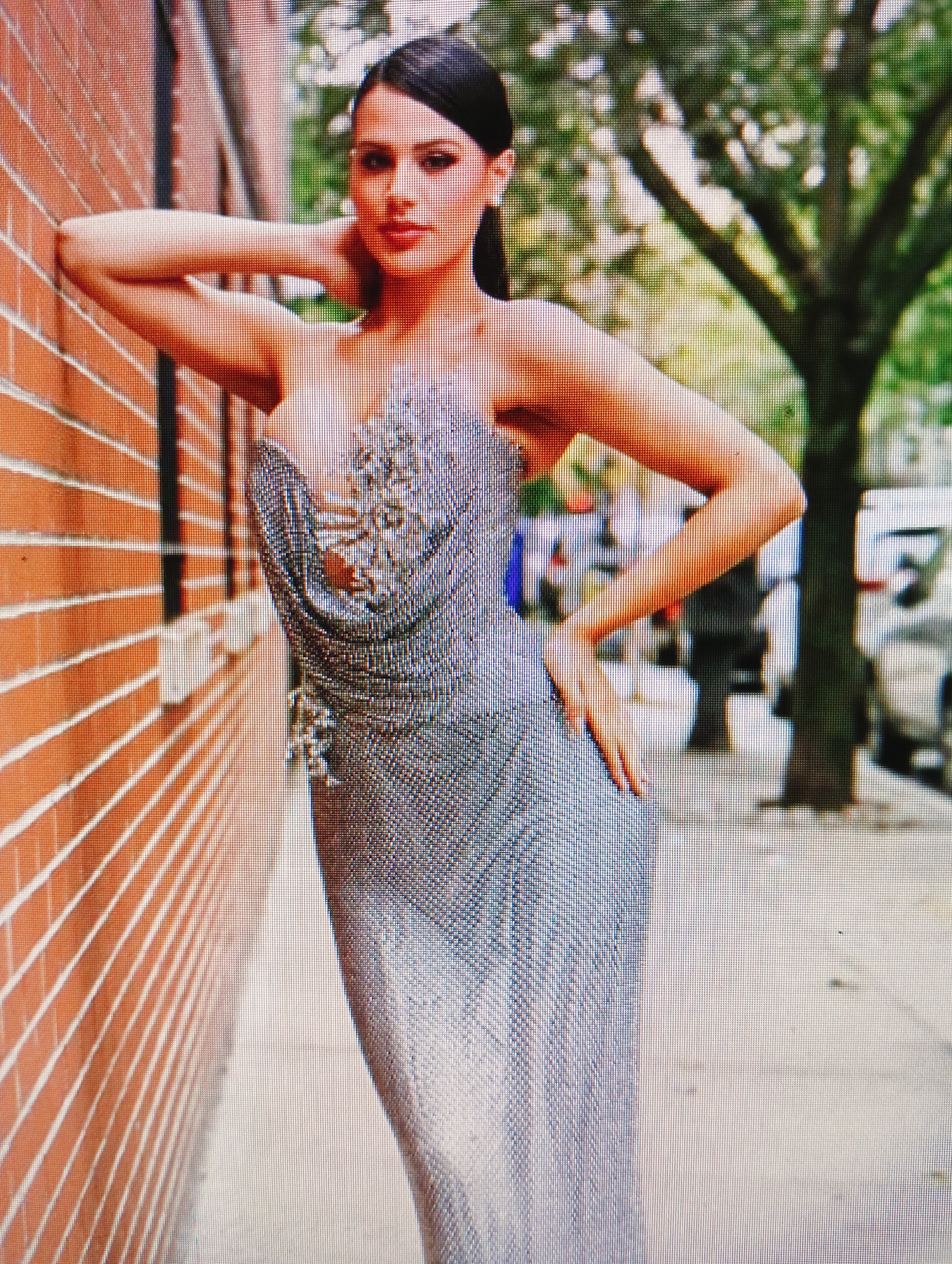 A full-body shot captures a woman leaning against a brick wall outdoors. She is wearing a strapless, shimmering silver gown adorned with intricate embellishments on the bodice. Her left arm is bent and resting on her head, while her right hand is placed on her hip. The woman has dark hair pulled back and is looking directly at the camera. The background consists of a brick wall on the left, a tree with green foliage in the center, and a sidewalk with blurred figures and vehicles on the right. The lighting suggests it is daytime. The image quality is low, indicating it may be a digital photograph of a screen.