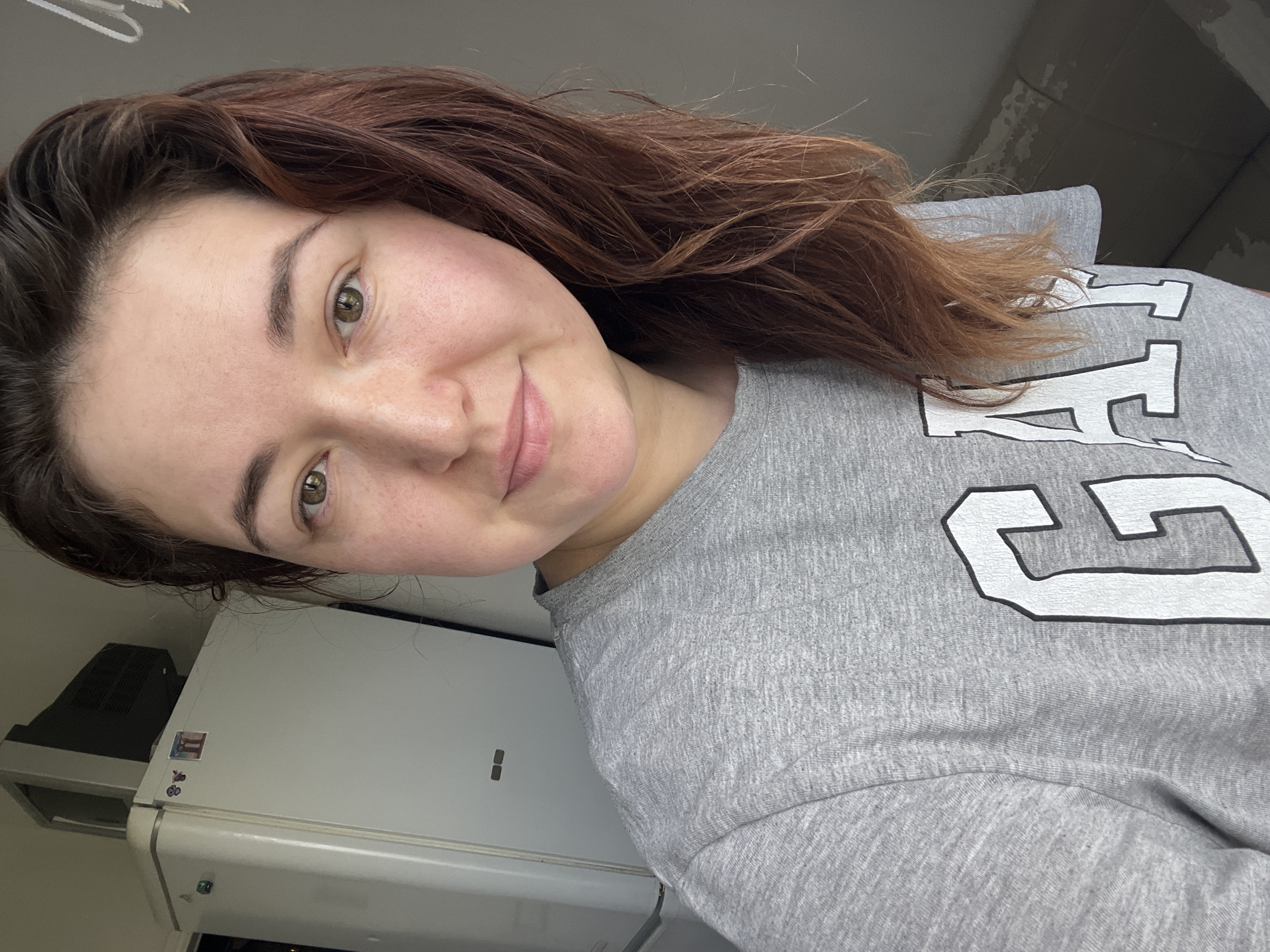 A woman with medium-length brown hair is taking a selfie indoors. She is wearing a grey t-shirt with the word "GAP" in large white letters. The background appears to be a residential setting, with a refrigerator and what looks like an old television set visible behind her. The lighting suggests it is daytime. There is no specific identifiable location in Chelyabinsk, Russia.