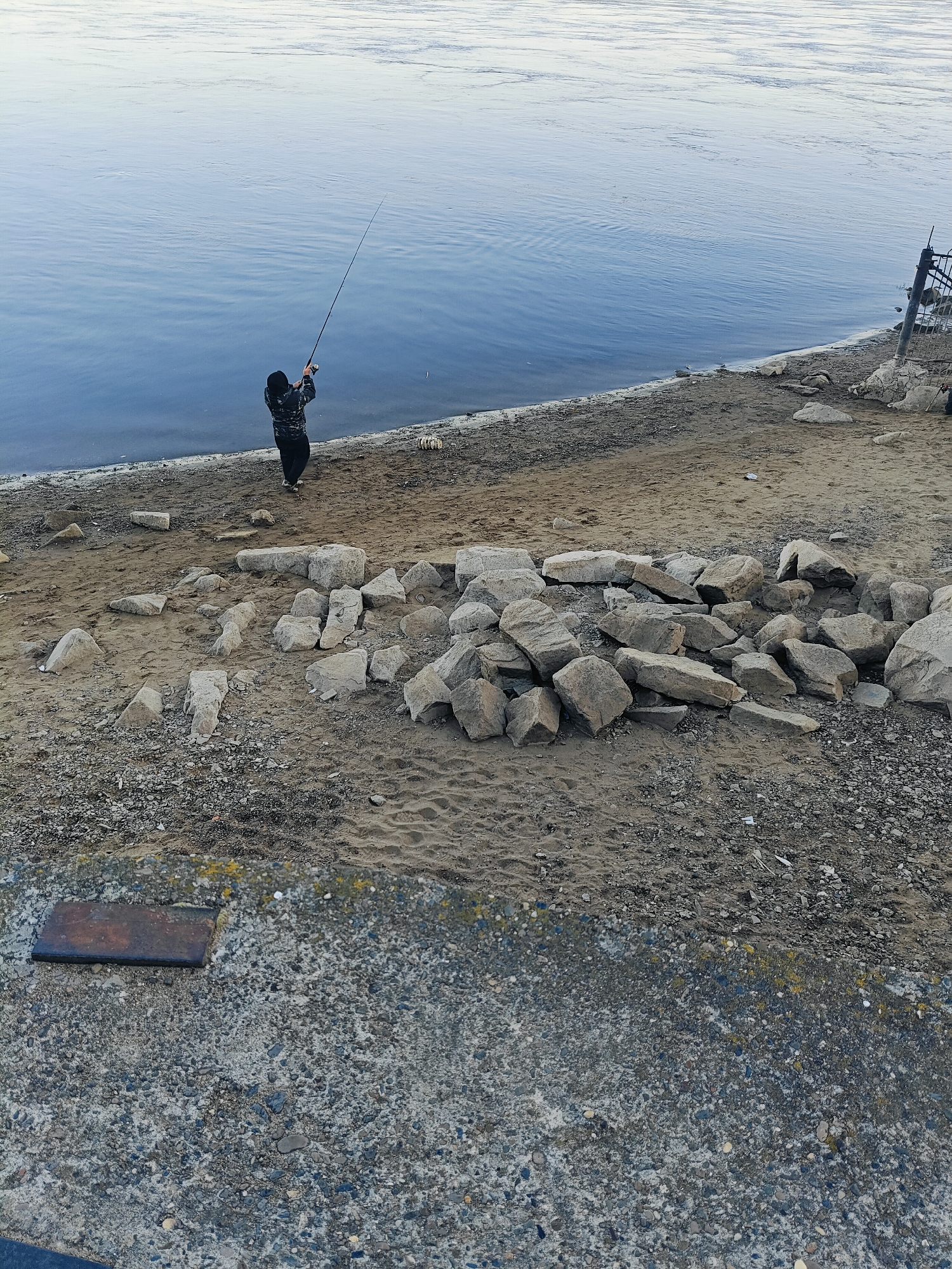 An elevated, full shot captures a solitary individual fishing on the sandy bank of a wide river in Chaikovsky, Russia. The river, a vast expanse of tranquil, pale blue water with subtle ripples, dominates the upper portion of the frame, reflecting a cloudy sky. The bank is a mix of sand and pebbles, with a noticeable pile of grey rocks forming a rough barrier in the mid-ground. The fisherman, dressed in dark clothing with a patterned jacket and a black hat, stands with their back to the camera, holding a long fishing rod extended towards the water. The foreground consists of a rough, grey concrete surface with a dark, rectangular metal plate embedded in it. A black metal fence structure is visible on the right side of the frame, near the riverbank. The overall scene suggests a quiet, perhaps early morning or late afternoon, fishing excursion.