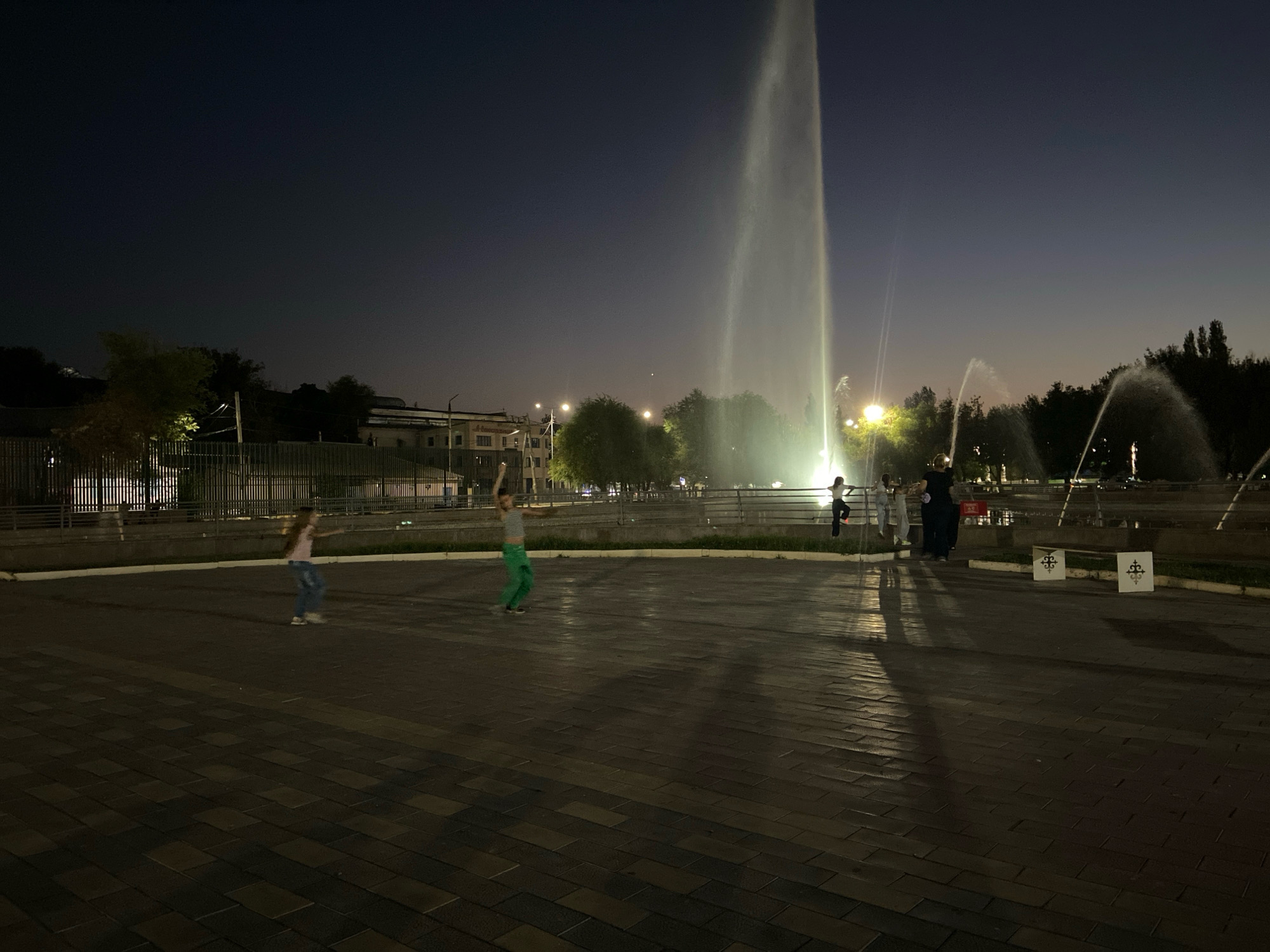 The image shows a night scene at the "Zvezda" fountain in Shymkent, Kazakhstan. A tall water jet from the central fountain dominates the background, illuminated by artificial lights, creating a dramatic visual effect against the dark sky. Several smaller fountains with arching water sprays are visible to the right.
In the foreground, two young girls are seen. One, on the left, is wearing a pink top and blue jeans and appears to be walking. The other girl, in the center, is wearing a striped top and green pants and is in motion, possibly dancing or playing. Further to the right, a group of four people, including at least two children and two adults, are standing near the fountain. They appear to be observing the fountain or interacting with each other. The ground is paved with dark rectangular tiles. The scene is characteristic of a public park or square, likely a popular spot for evening recreation.