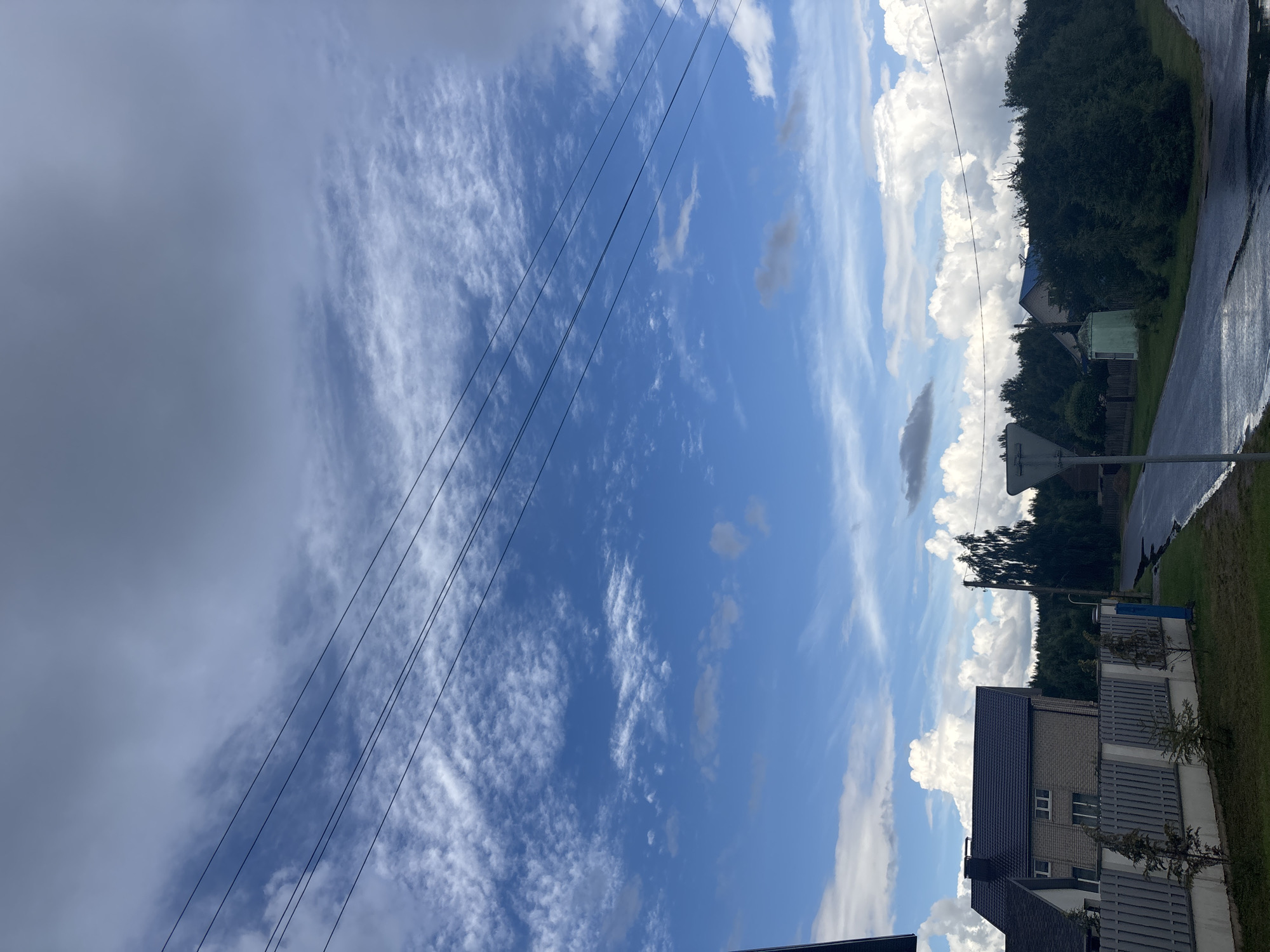 This image depicts a rural scene in the vicinity of Капыльскі сельскі Савет, Belarus. The sky is partly cloudy with a mix of white cumulus clouds and darker, more expansive grey clouds. Several power lines are strung across the upper portion of the frame. In the foreground, a paved road, wet from recent rain, curves to the right. To the left of the road, a grey fence borders a property with a modern house featuring a dark-tiled roof and brick exterior. Small, conifer-like trees are planted along the fence. A street sign is visible near the center of the image. Dense green trees form a backdrop behind the road and the houses. The overall impression is a quiet, post-rain day in a suburban or rural Belarusian landscape.